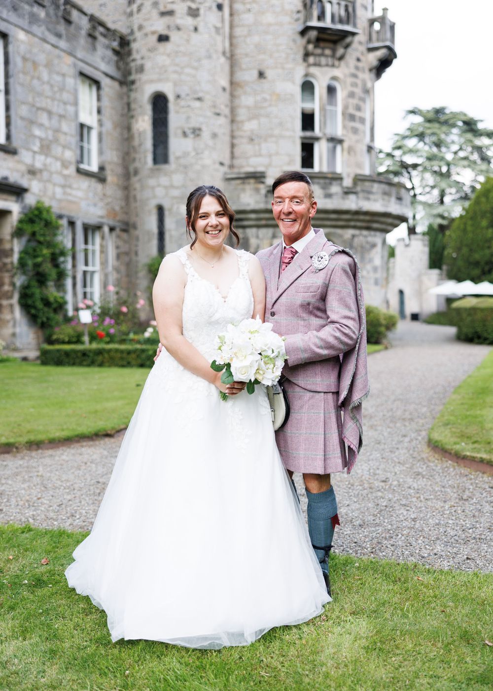 Bride in white gown and groom in pink tartan kilt pose together outside a Scottish castle on their wedding day.