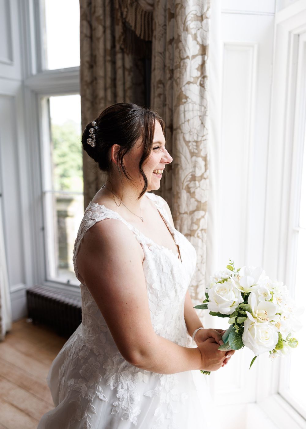 Smiling bride in lace wedding dress holding white rose bouquet standing by window in elegant venue.