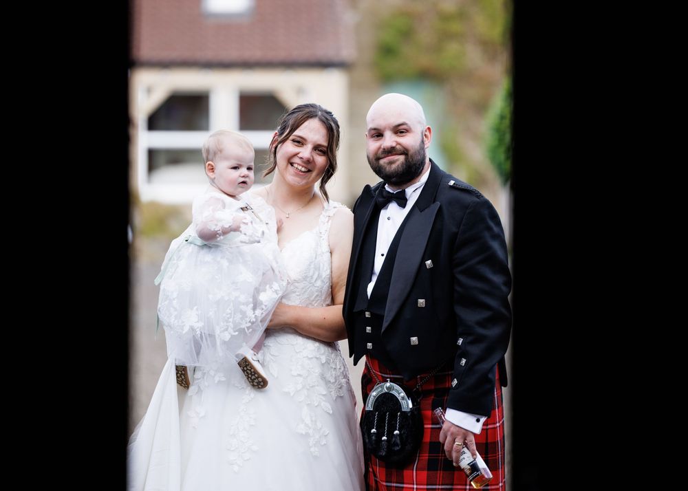 Smiling bride and groom in Scottish kilt holding baby in white christening gown outdoors.