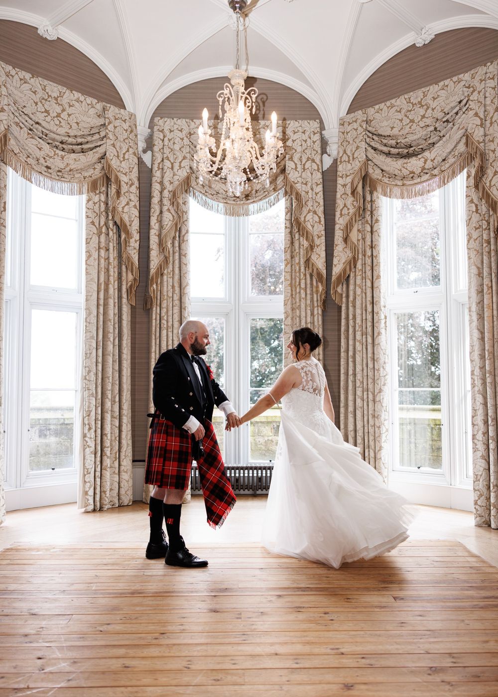 Bride and groom share a dance in elegant room with arched windows, chandelier, and ornate curtains.