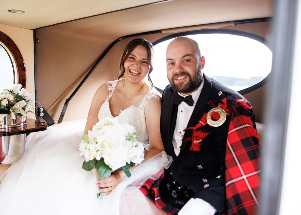Smiling newlyweds sit in vintage car, groom in red tartan kilt, bride holding white floral bouquet.
