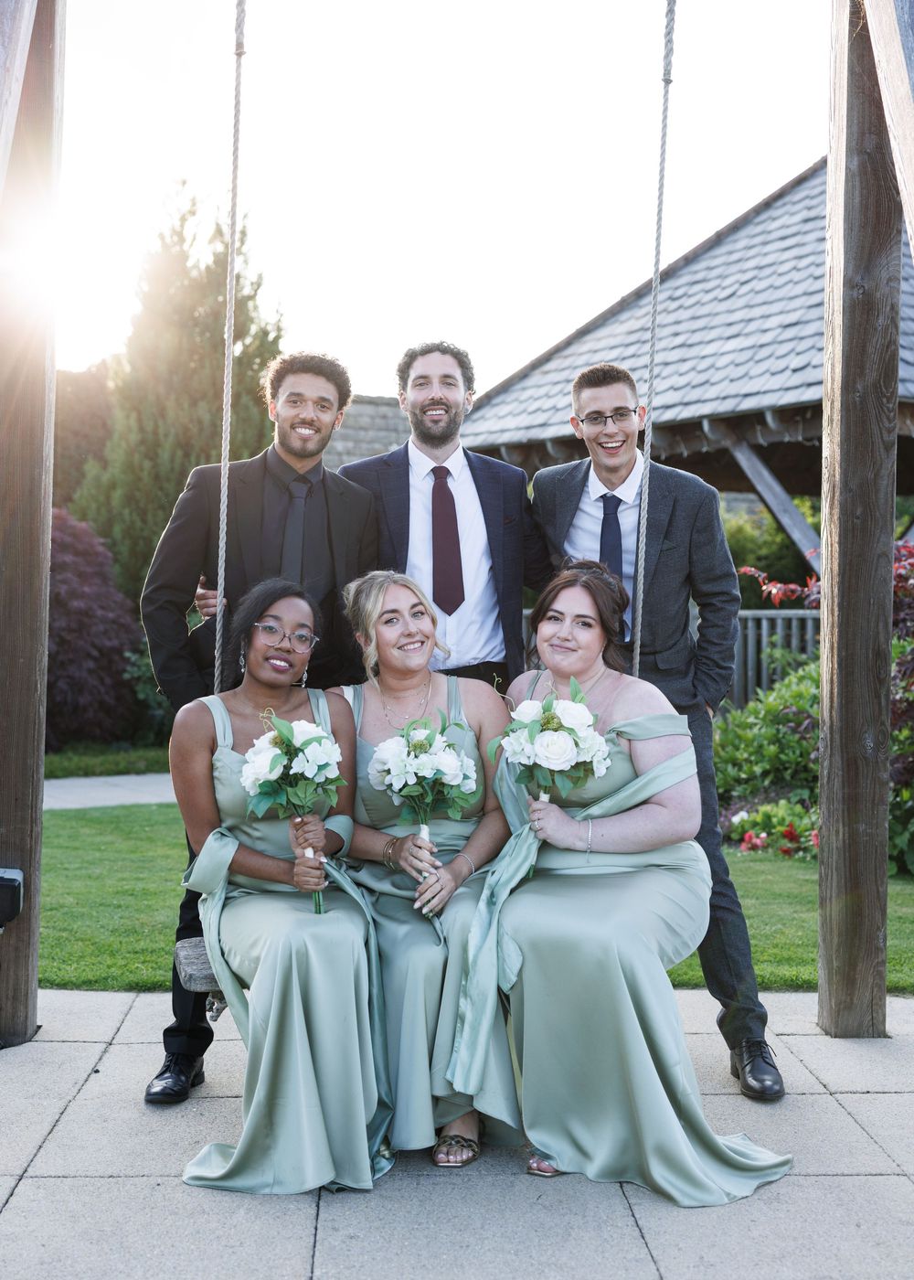 Three bridesmaids in sage green gowns holding white bouquets sit on a swing, with three groomsmen standing behind them.