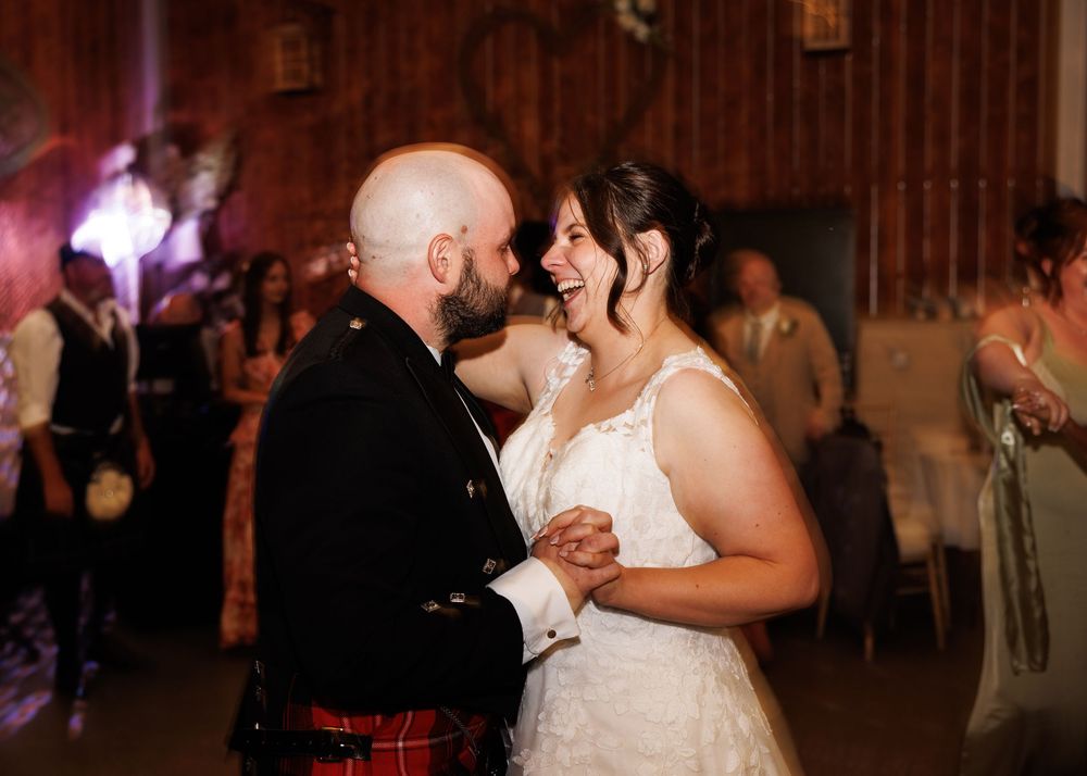 Bride and groom sharing first dance, groom in red tartan kilt, bride in lace gown, smiling joyfully at wedding reception.