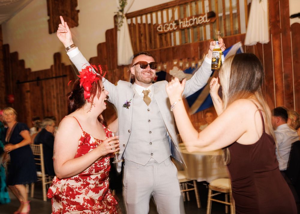 Groom in light grey suit celebrating at wedding reception, dancing with guests in a rustic barn venue.