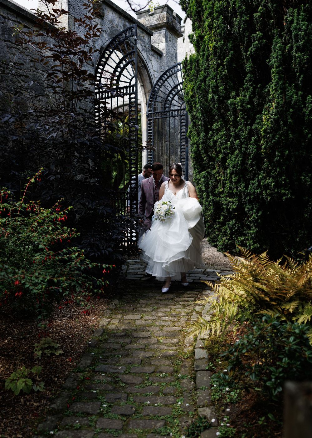 Bride in white gown walks through ornate iron garden gate on cobblestone path surrounded by lush greenery.