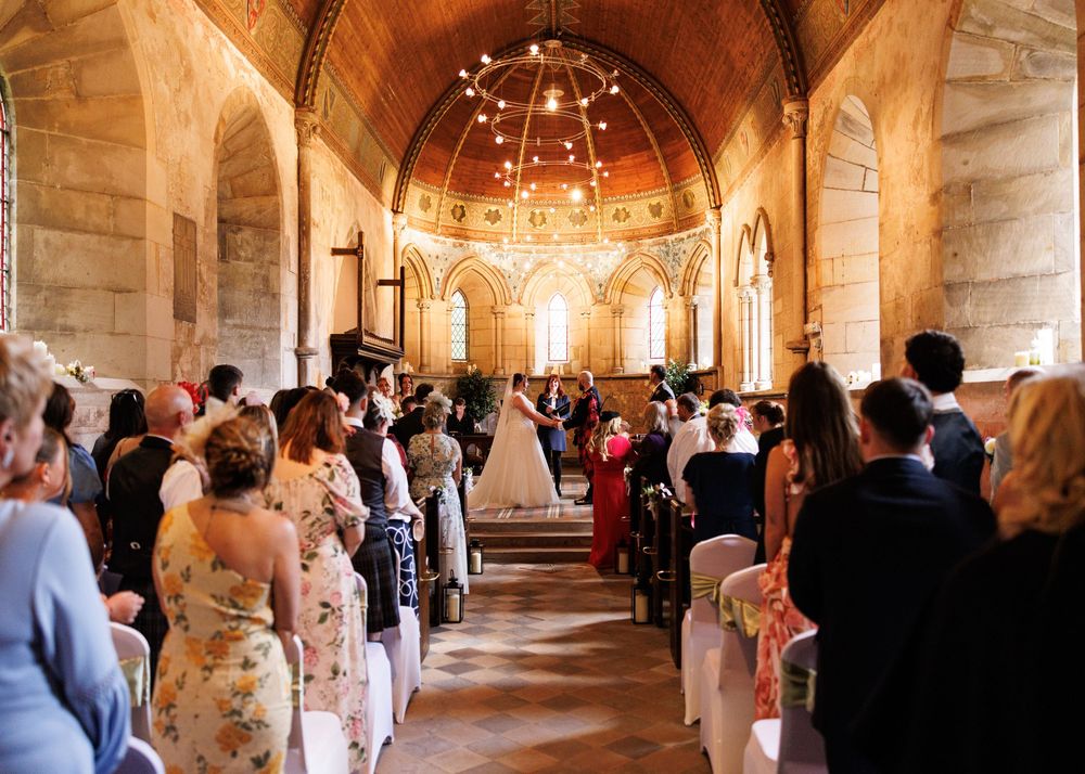 Wedding ceremony inside a stunning stone church with vaulted ceiling, guests standing as bride walks down the aisle.
