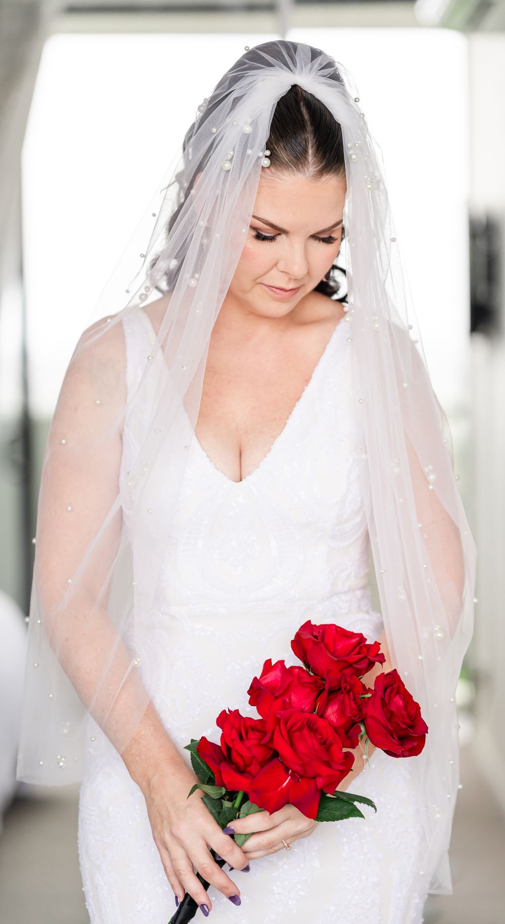 Bride in white dress and veil holds vibrant red roses during wedding photoshoot.