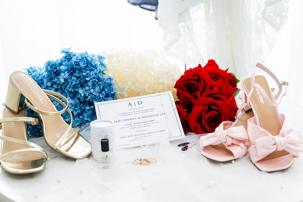 Wedding accessories including shoes, rings, flowers and invitations arranged on white surface.