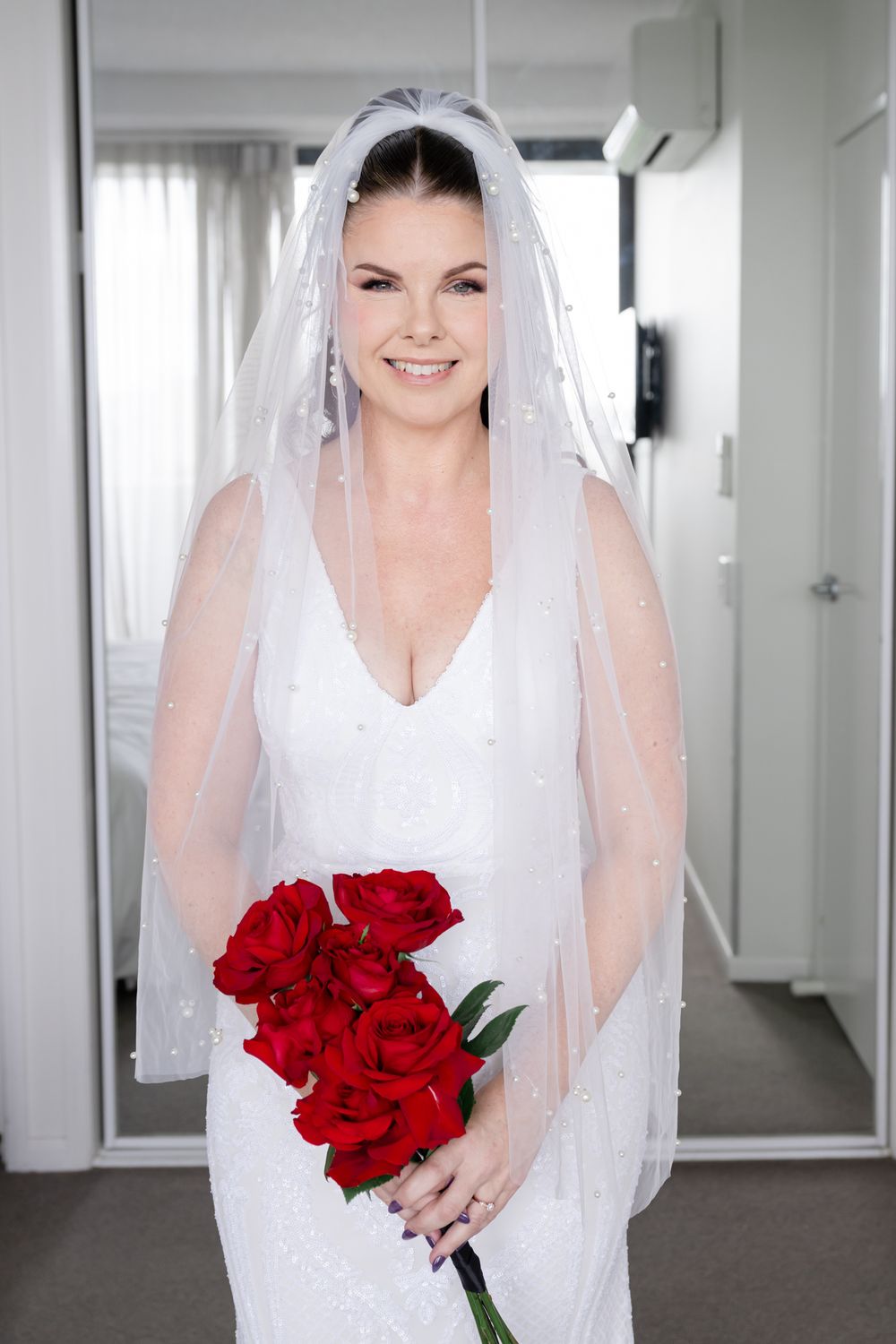 Bride in white wedding dress holding red rose bouquet in bright indoor setting.