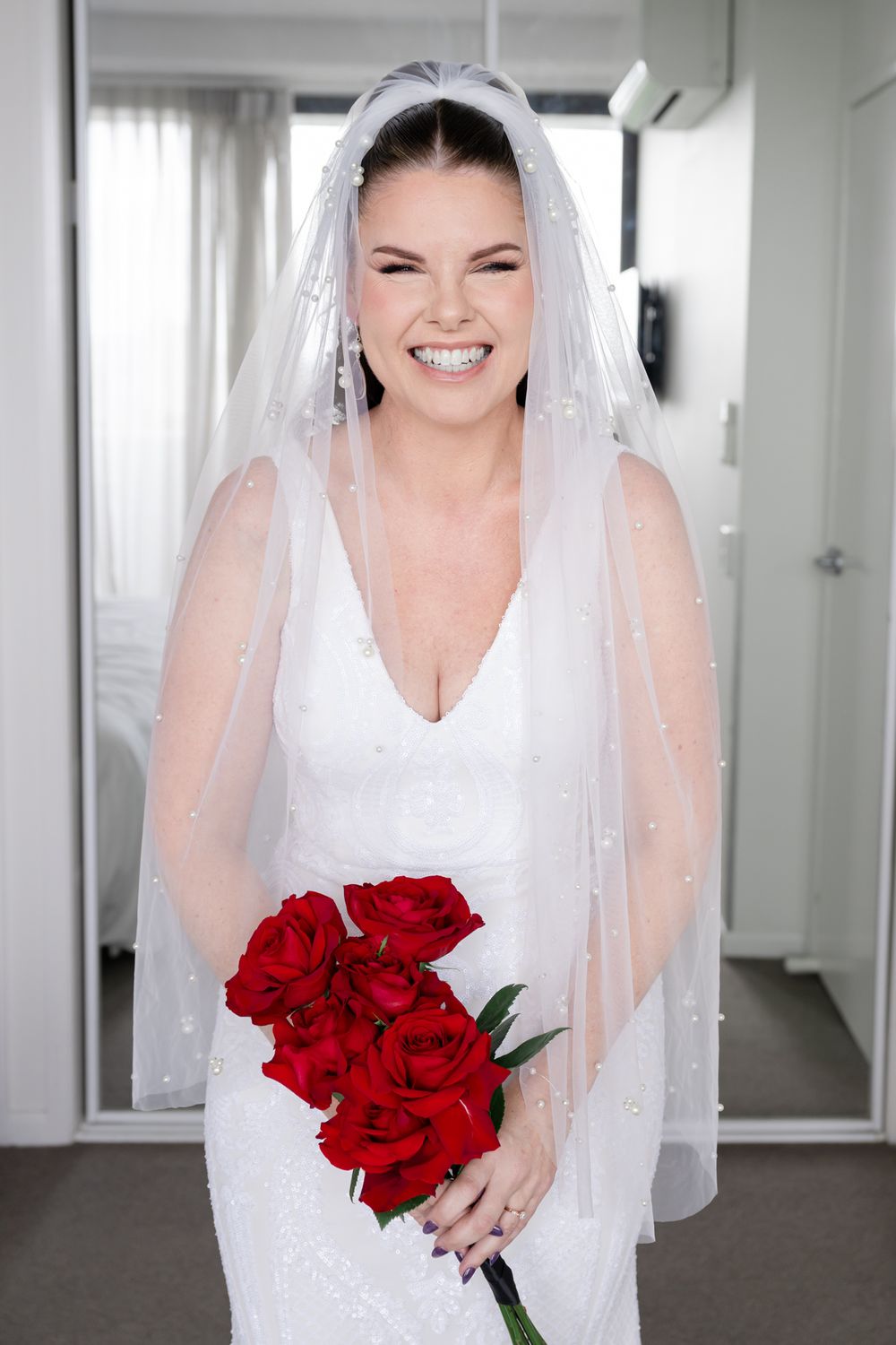 A bride in a white wedding dress and veil holds a bouquet of red roses while standing in a bright room.