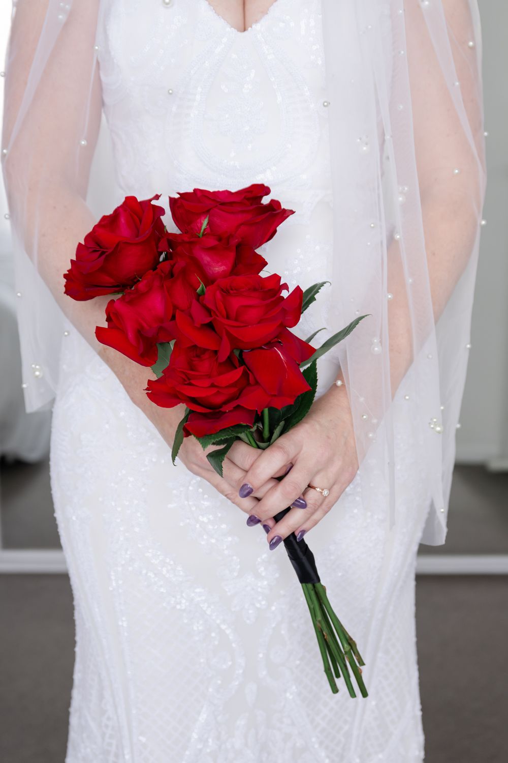 Close-up of a bridal bouquet with vibrant red roses against a white wedding dress and sheer veil.