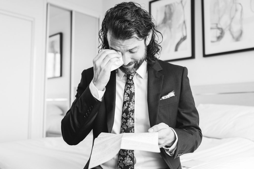 Black and white photo of someone in a suit and floral tie reading a document while touching their face in distress.
