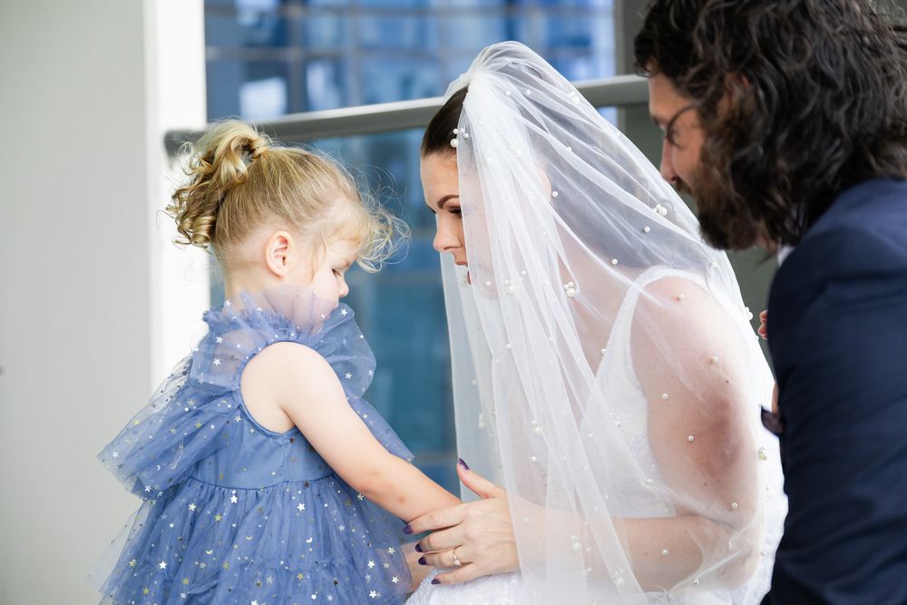 A bride in a white wedding dress and veil embraces a young child in a blue dress during a tender moment.