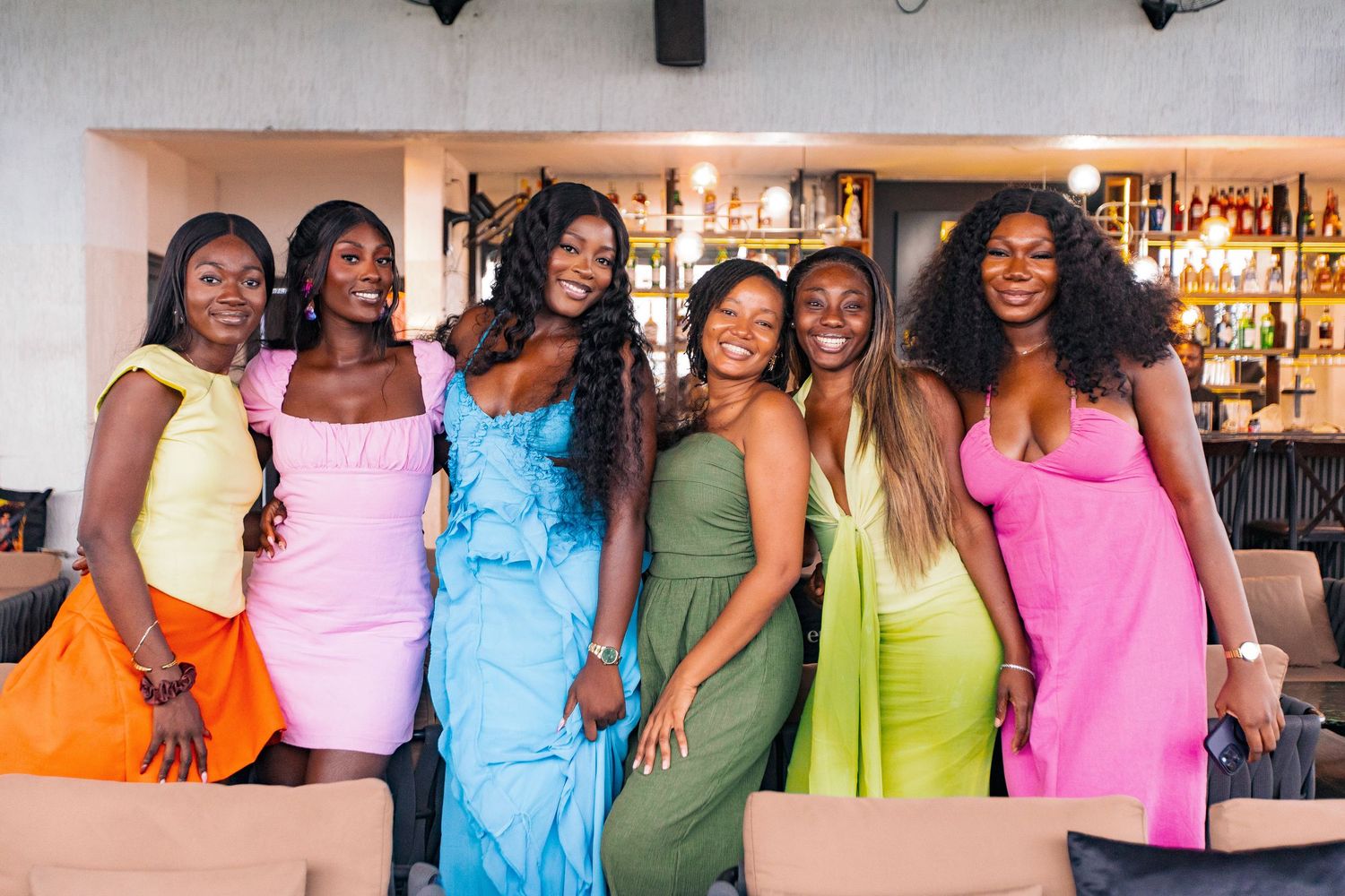 A group wearing vibrant formal dresses in yellow, pink, blue, green and other colors pose together at a bar.