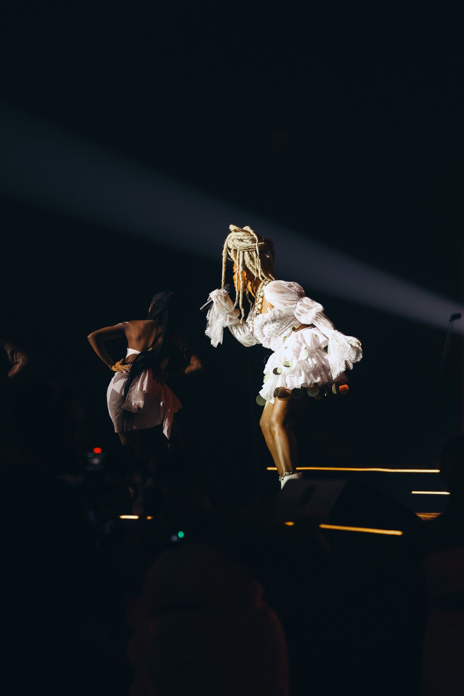 Two dancers in white costumes perform energetically under dramatic spotlights on a dark stage.