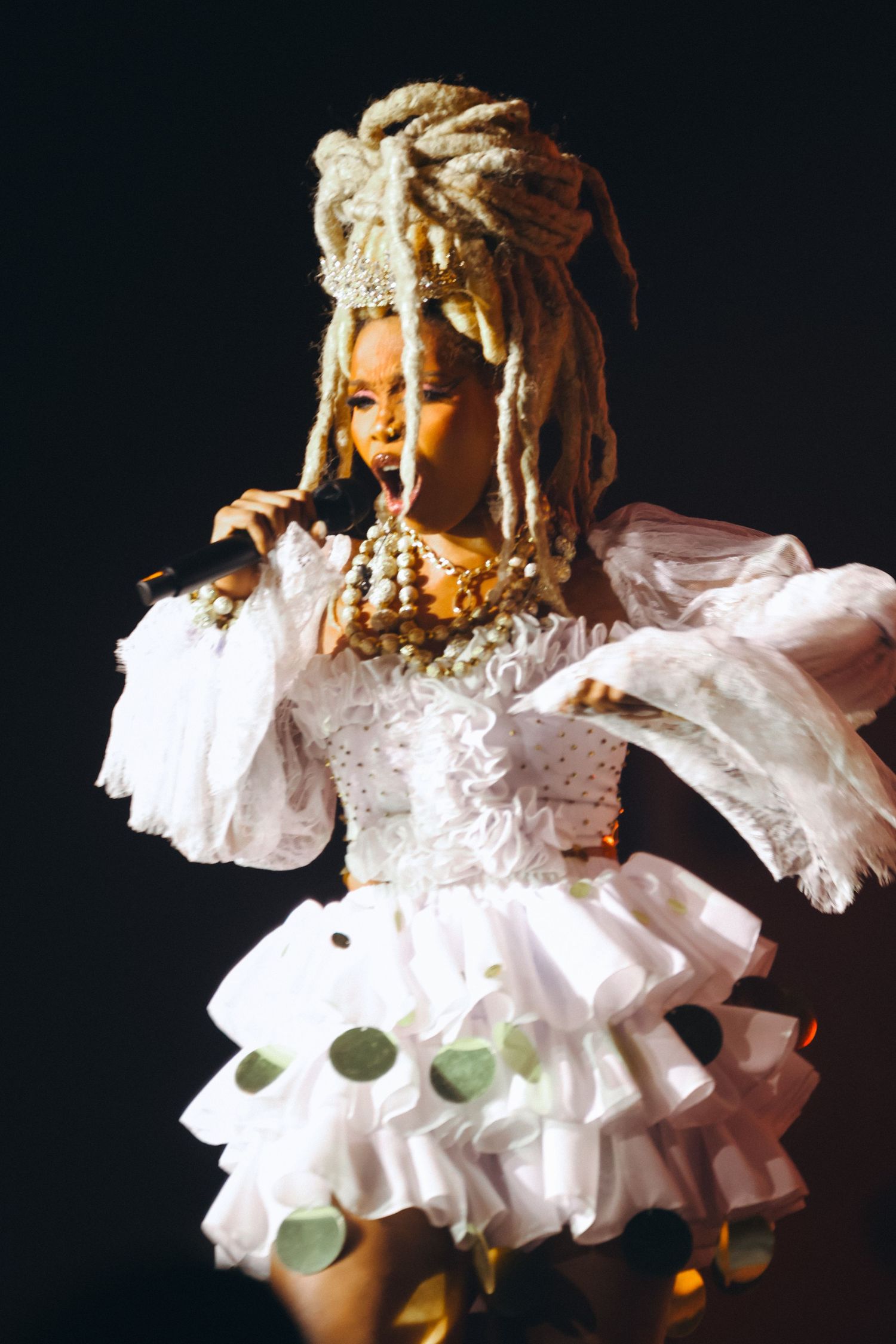 Female performer in white ruffled dress, gold jewelry, and dreadlocks headpiece singing on stage.