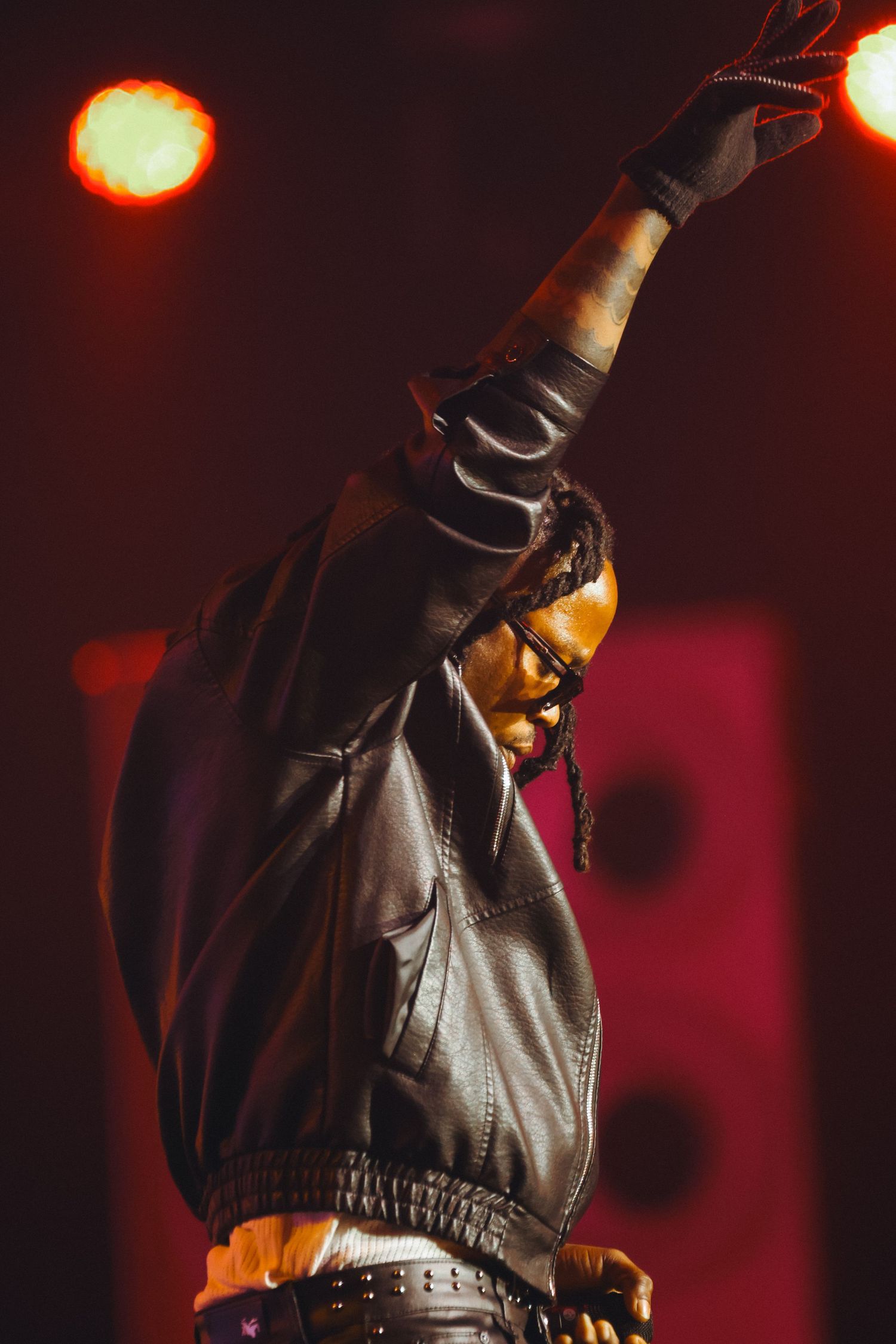 A performer in black leather raises their fist dramatically under red stage lighting at a live concert.