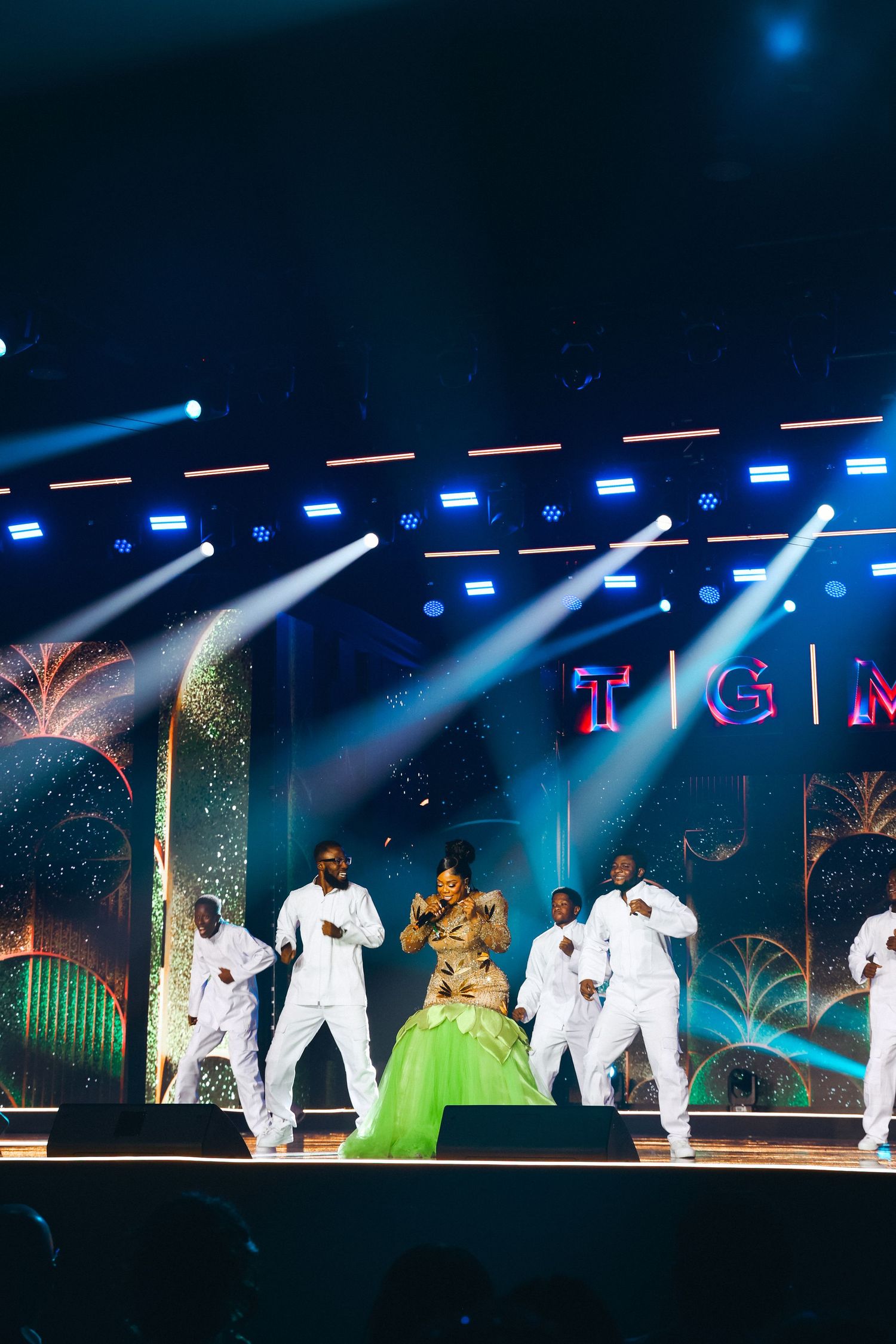 Female artist in gold and green gown performs with dancers in white at Ghana Music Awards.