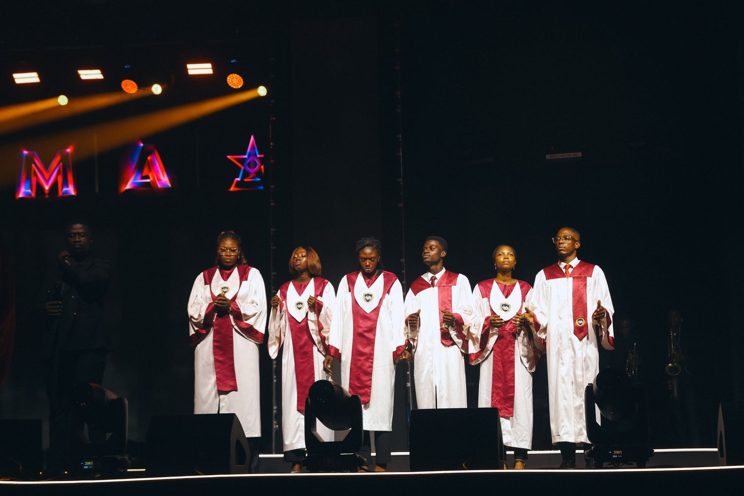 Gospel choir in white and red robes performing on a festive stage with colorful star decorations and dramatic lighting.