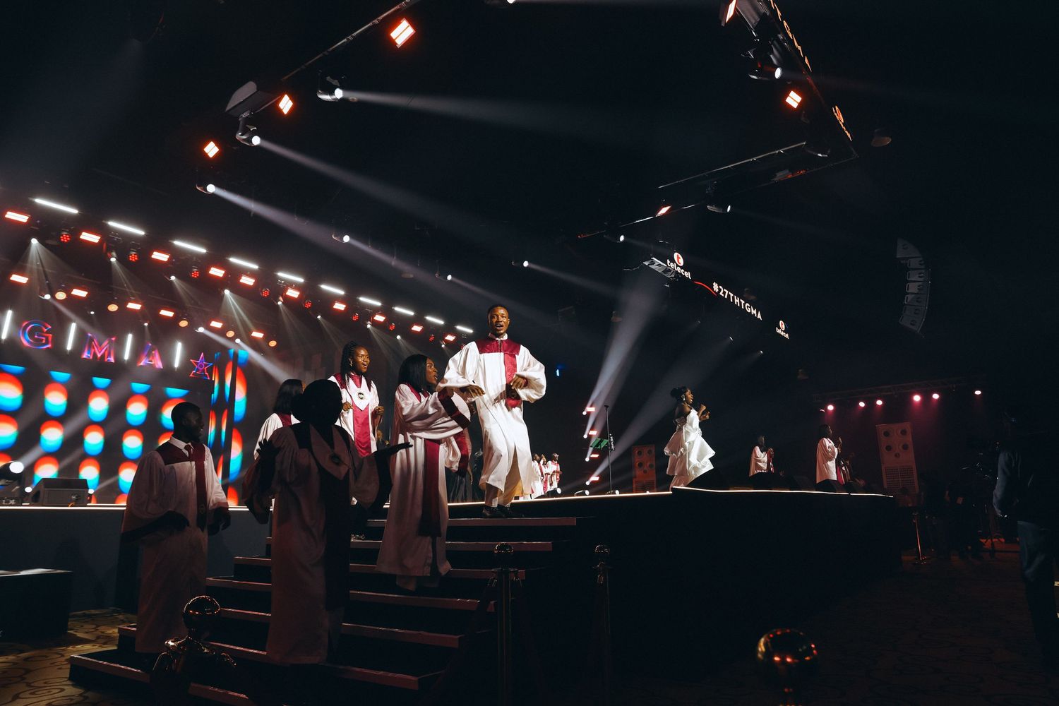 Performers on stage in white outfits under colorful spotlights at live show.