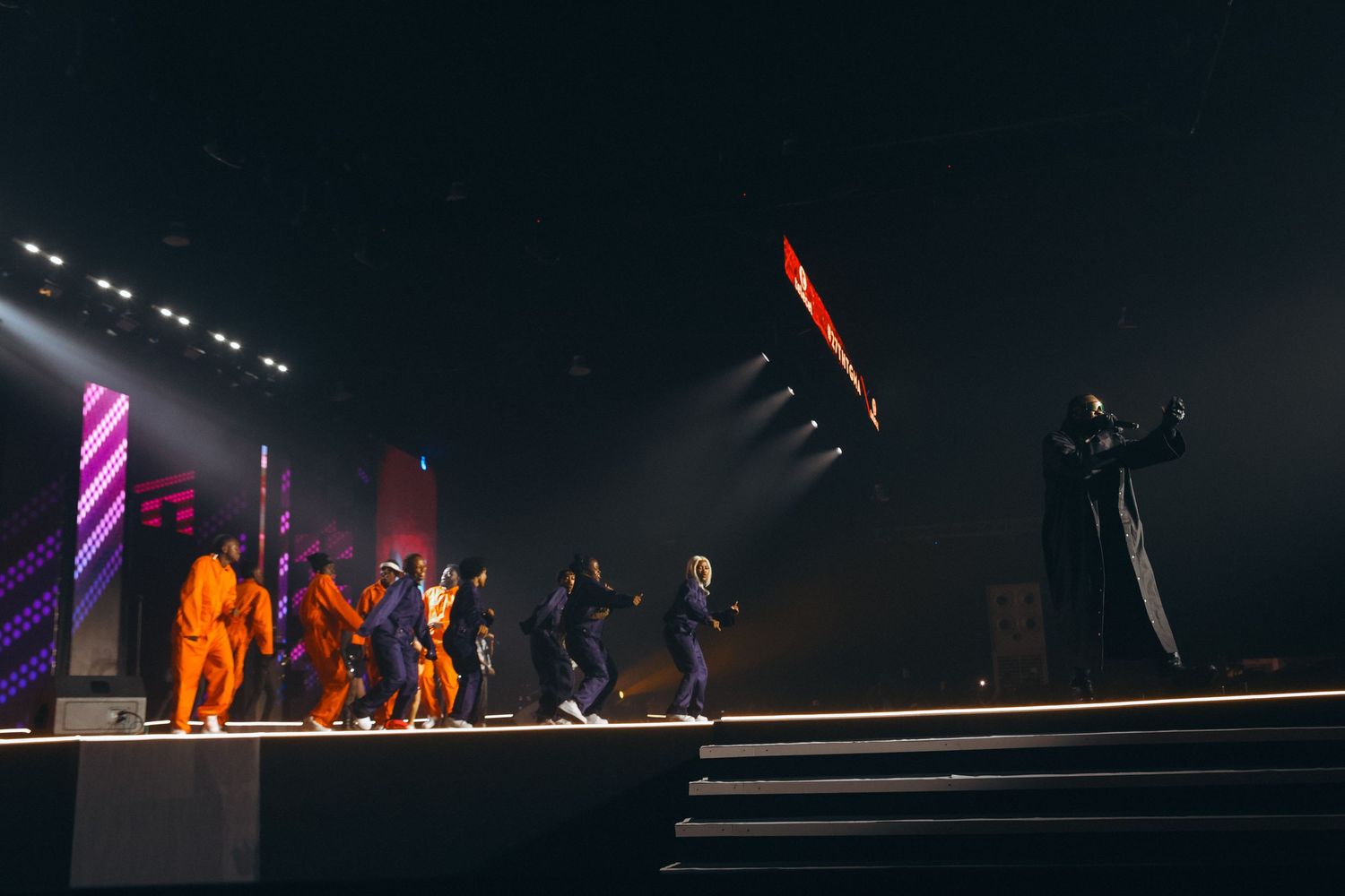 Large group of performers on a dark concert stage with colorful diagonal light beams during live show.