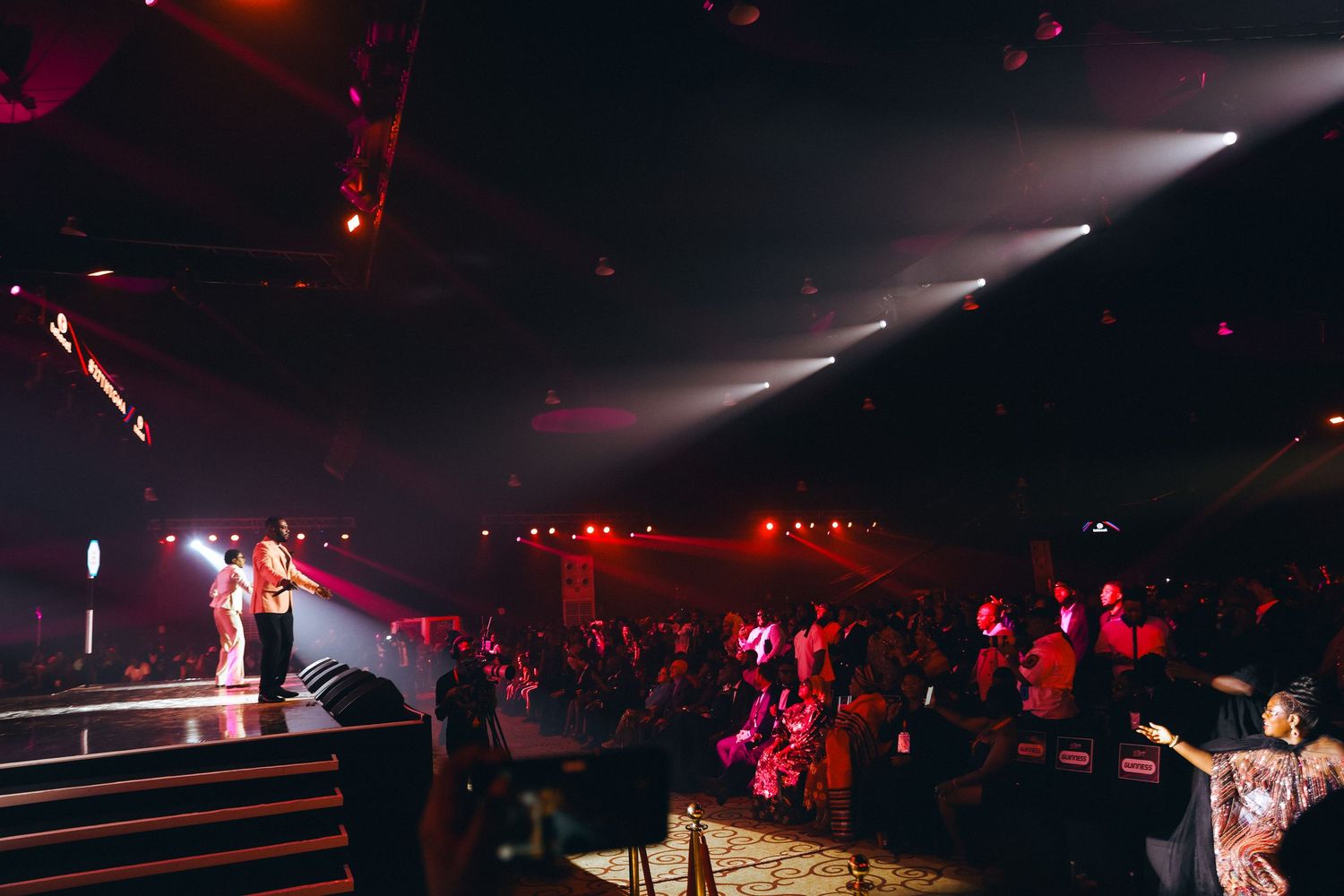A live concert performance with a crowd gathered under red stage lighting in a large indoor venue.