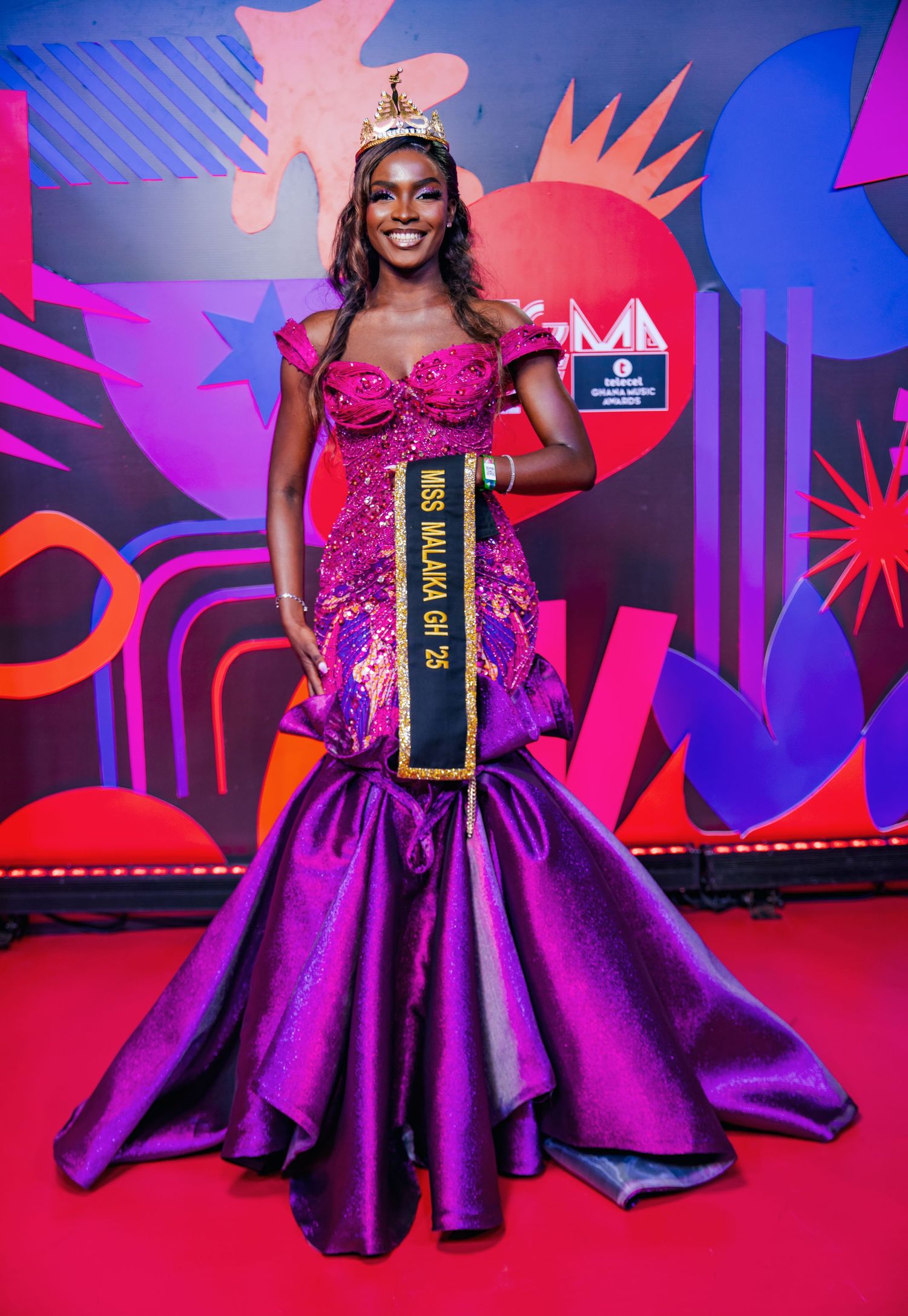 Miss Malaika Ghana 25 poses in purple mermaid gown and crown on red carpet at Ghana Music Awards.