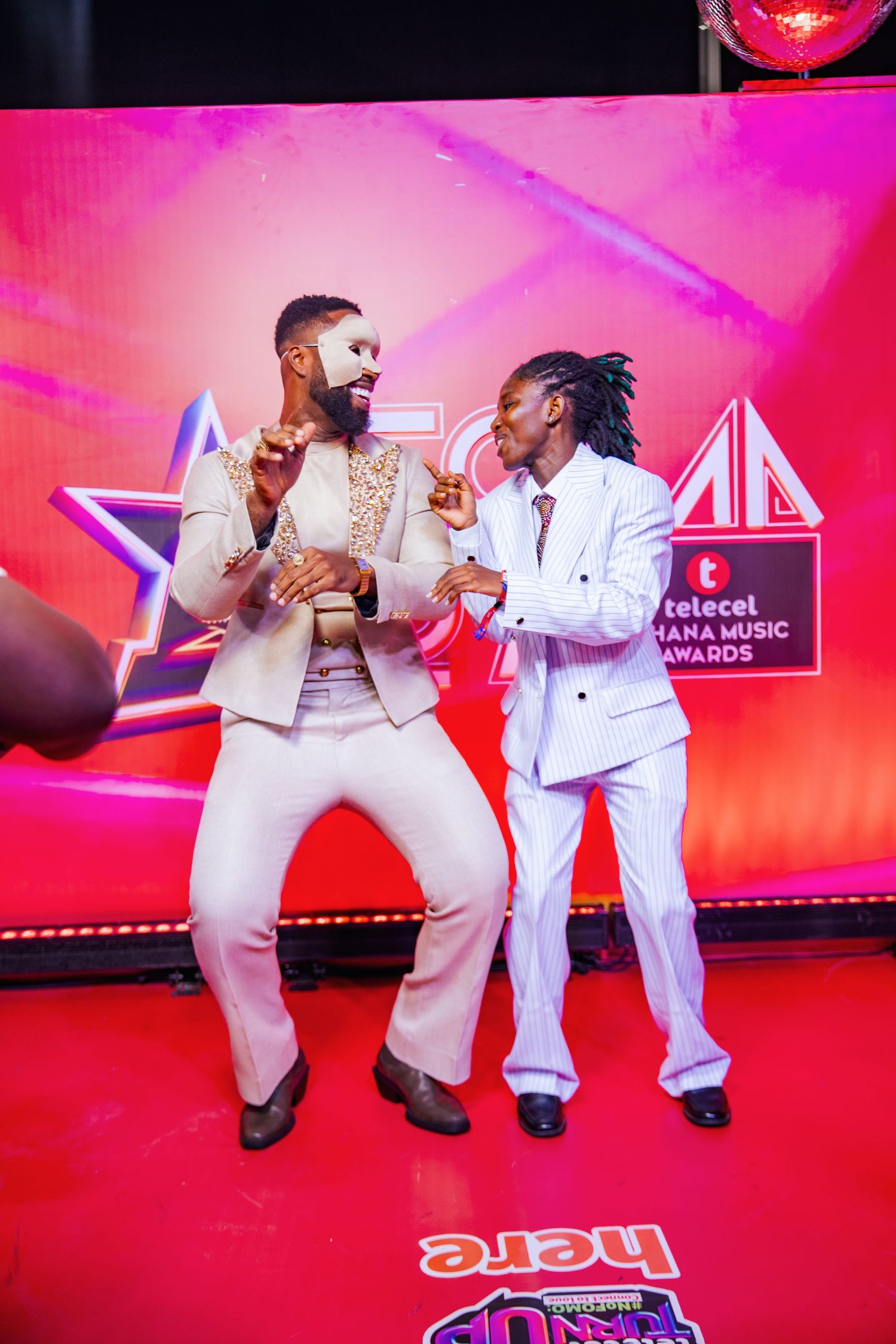 Two male artists in white suits dance and pose on Telecel Ghana Music Awards red carpet.