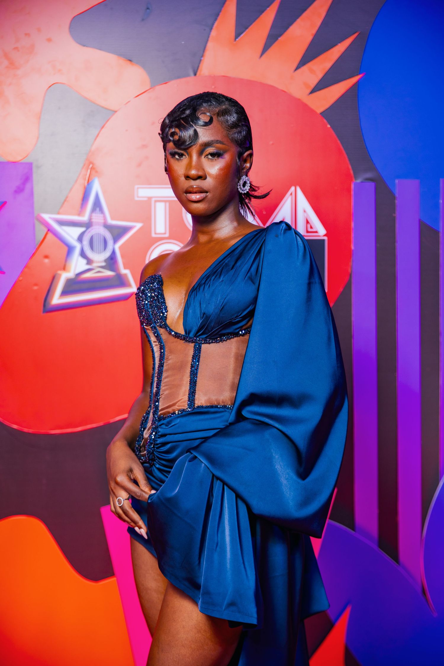 Woman in blue satin cutout mini dress with dramatic sleeves poses at Ghana Music Awards ceremony backdrop.