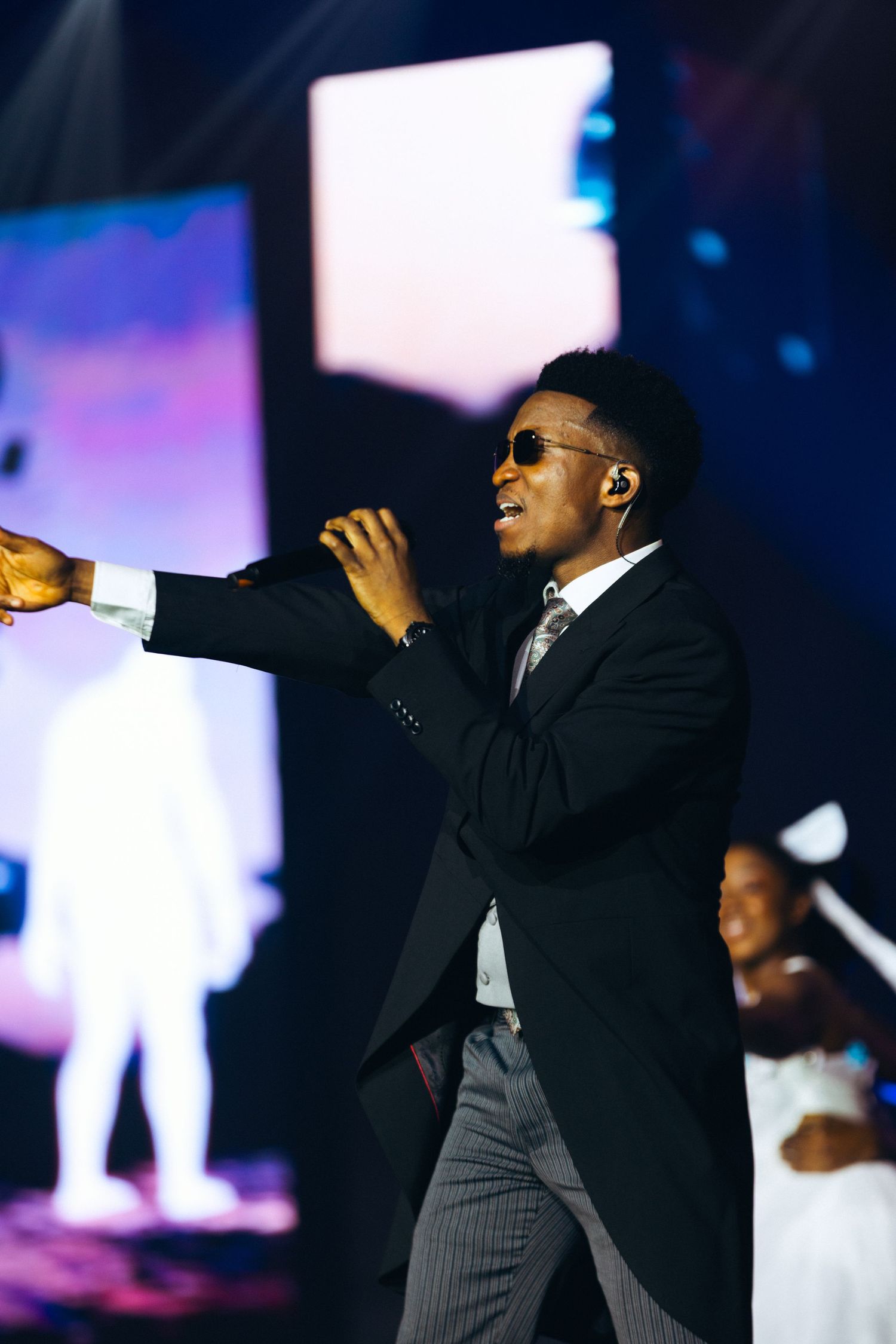 A stylish man in a black suit and sunglasses performs energetically on a colorful concert stage.