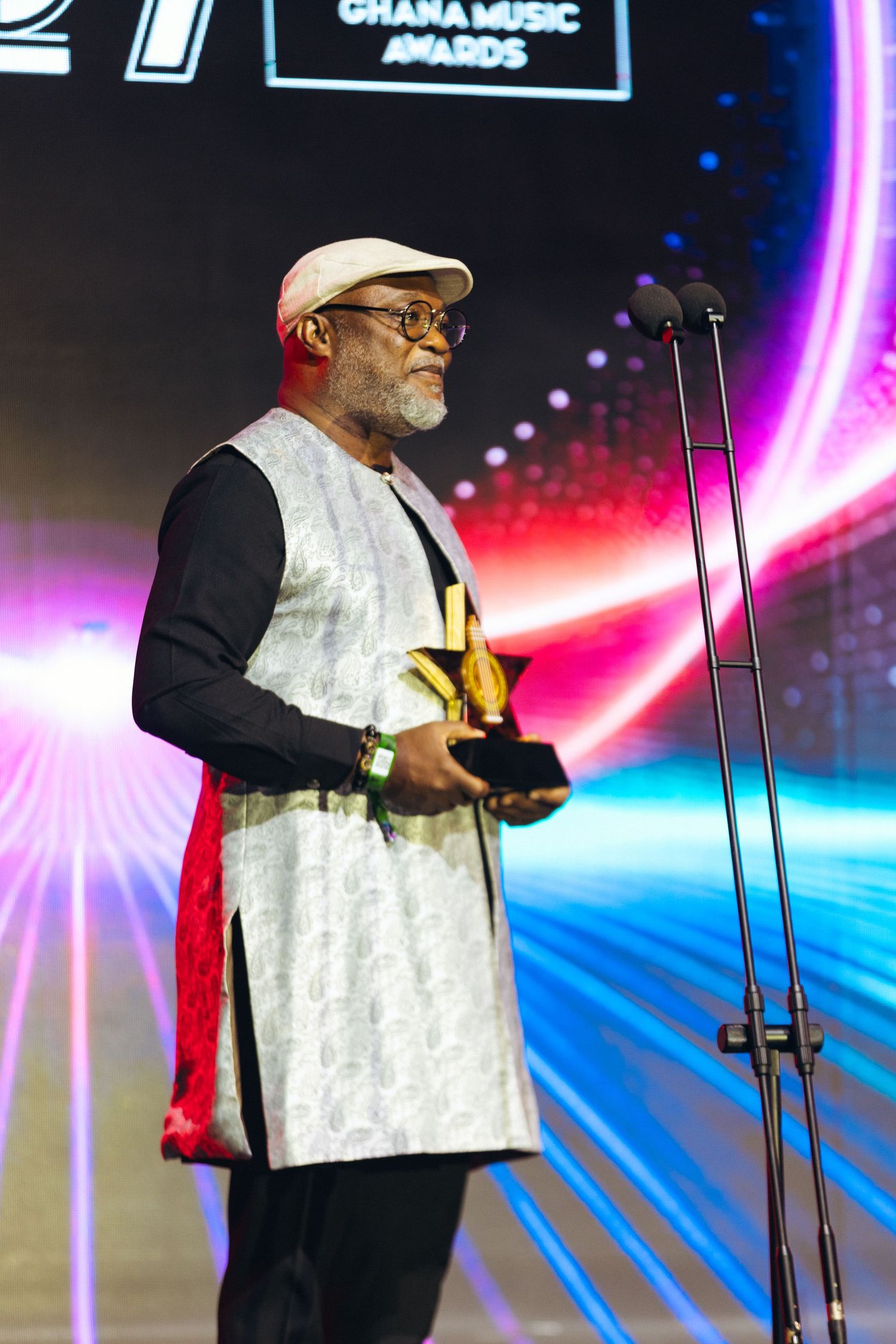 Older man in white embroidered vest accepts award at 27th Ghana Music Awards ceremony on stage.