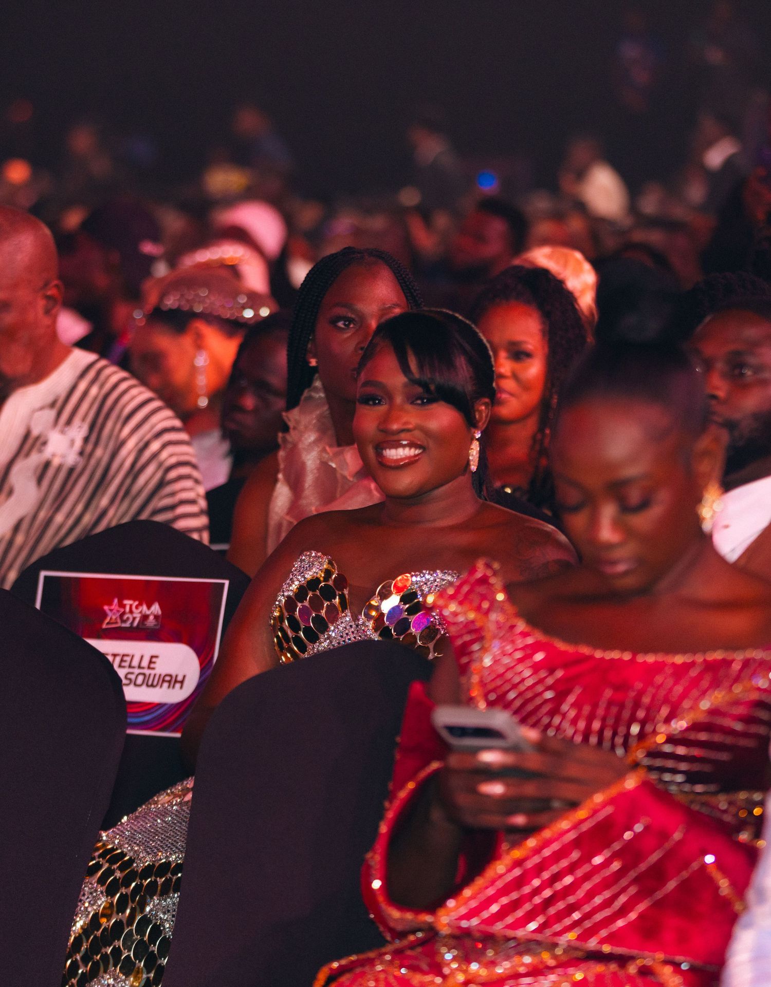 A smiling woman in a sparkling strapless dress enjoys a live awards show in the audience.