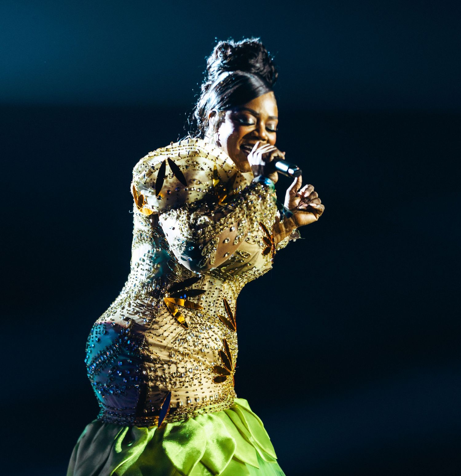 Female performer in gold sequin top and green skirt sings on stage at music awards show.