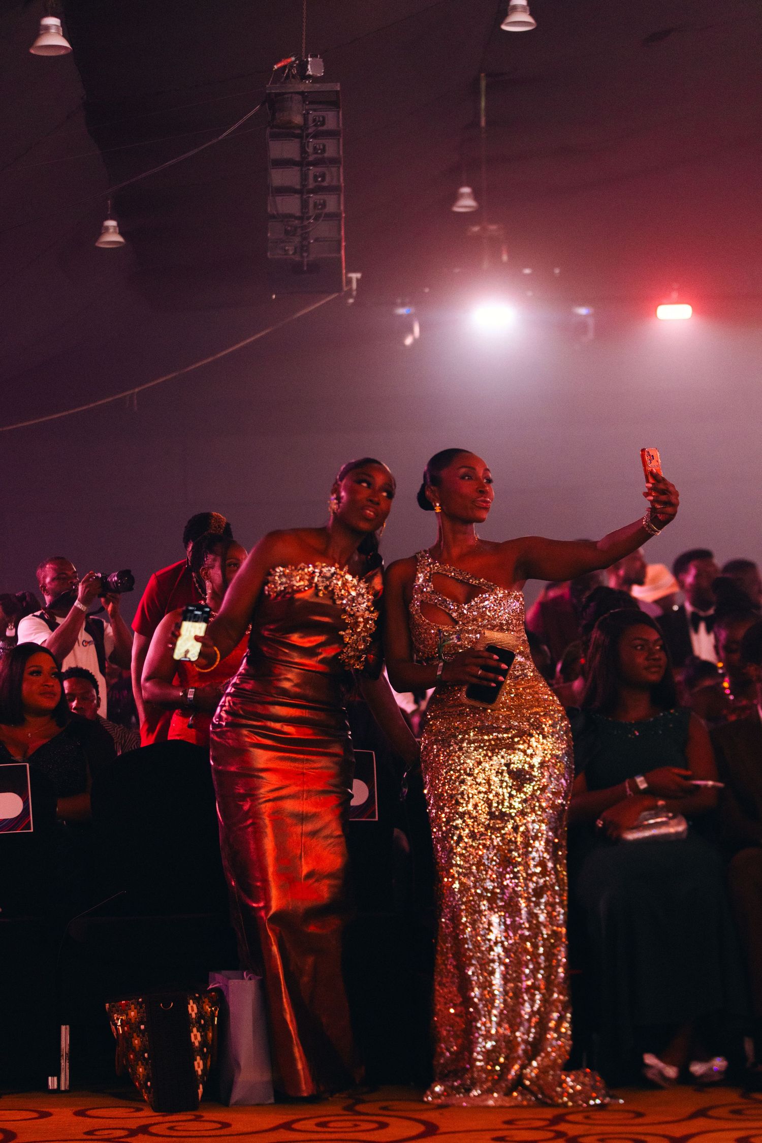 Two women in sequin gowns taking selfie at glamorous awards ceremony.