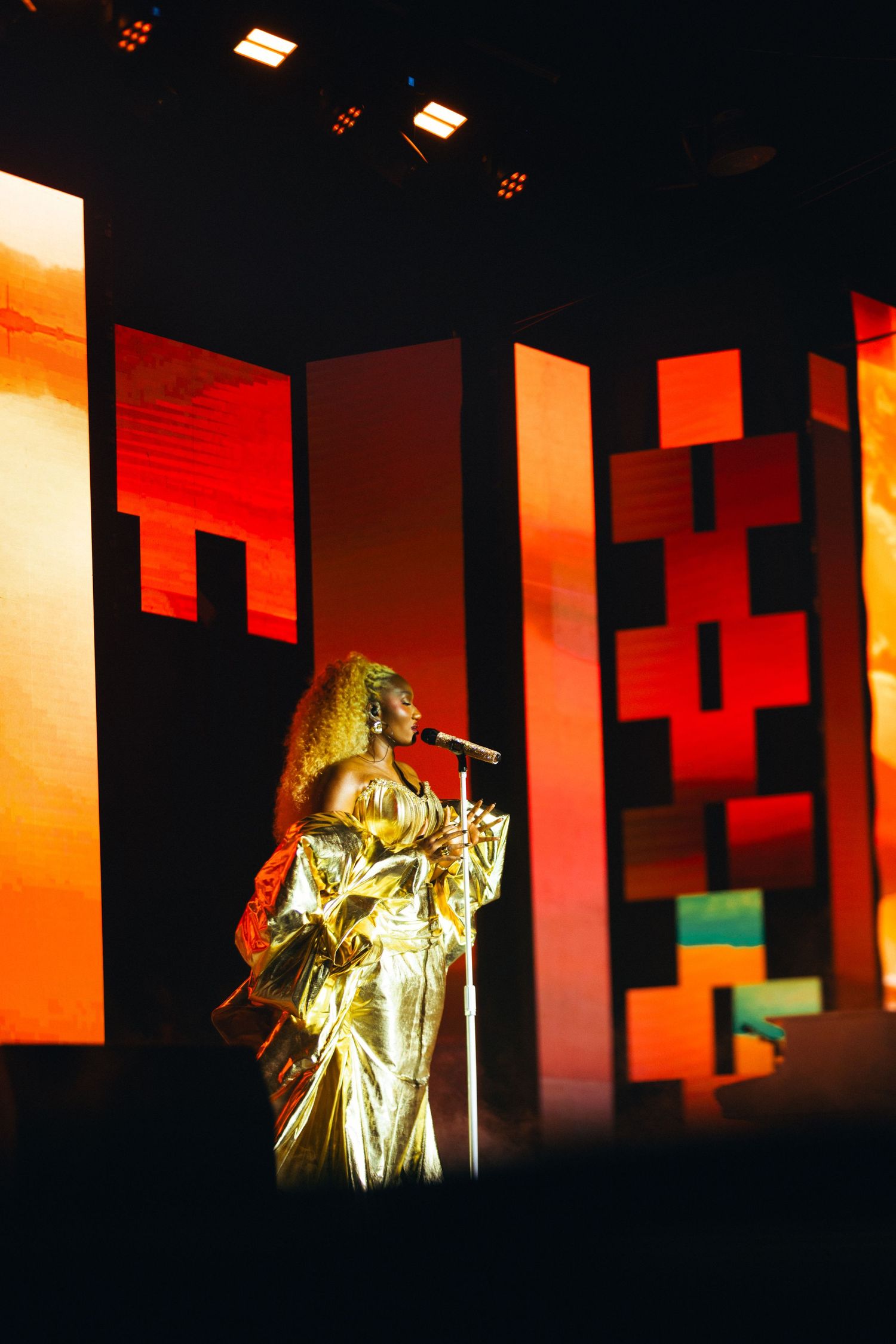 A performer in a gold gown sings at a microphone on a dramatic stage with red and orange geometric backdrops.