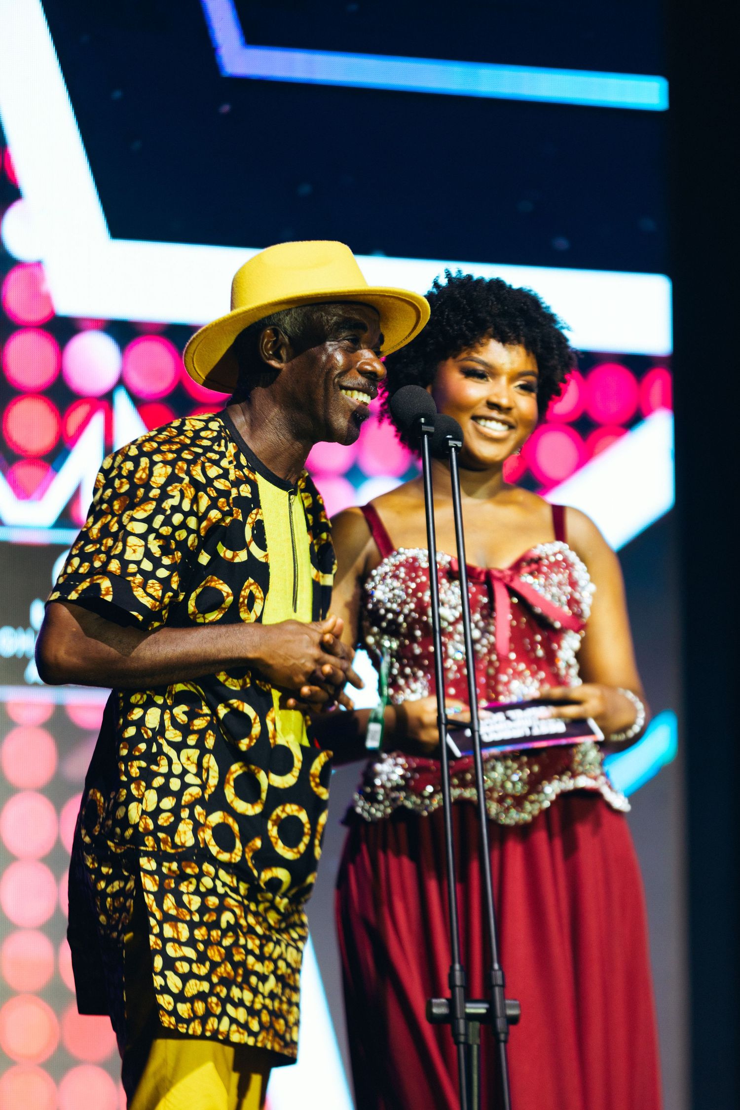 Man in yellow hat and African print outfit presents award with woman in red gown at Ghana Music Awards.