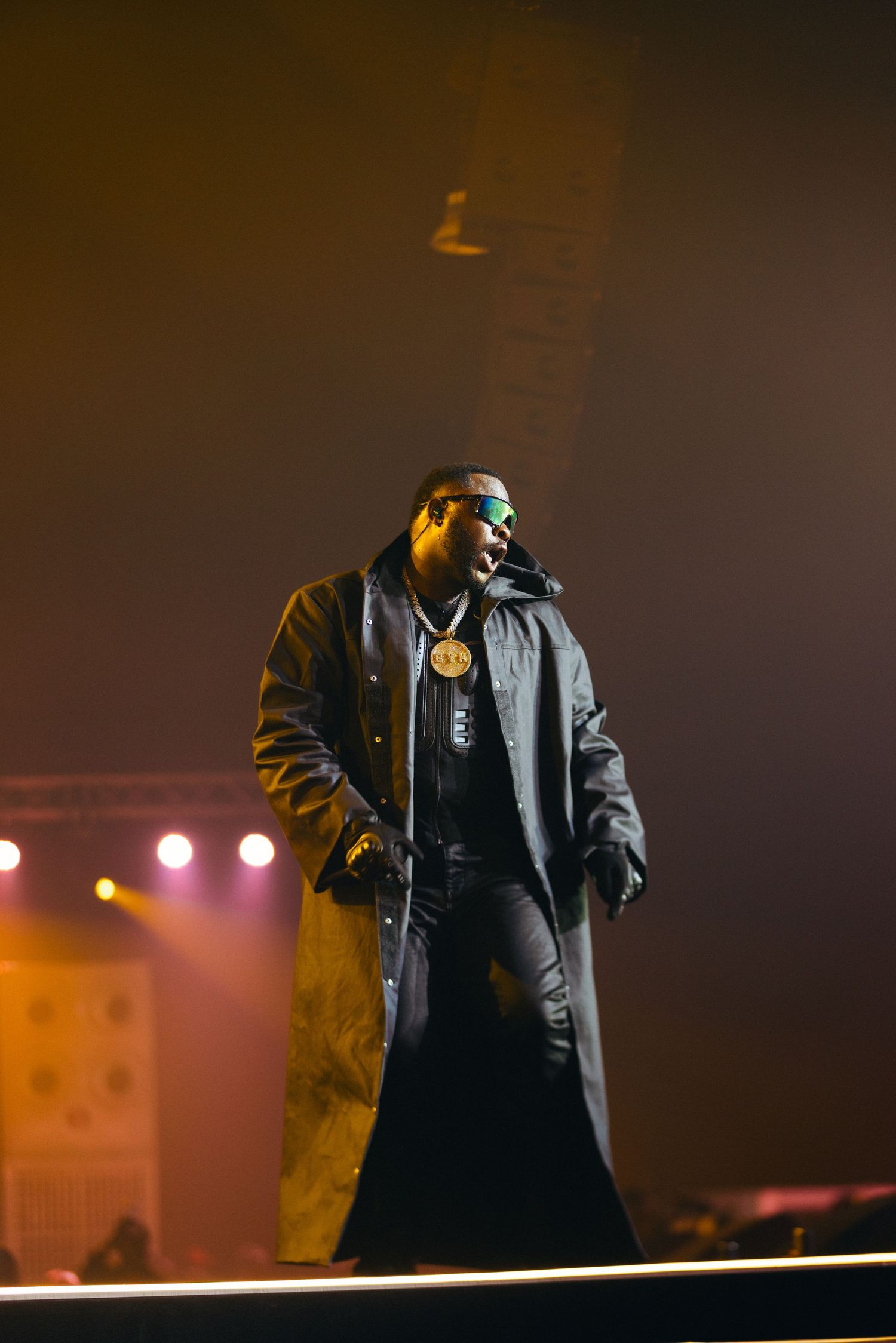 Performer in long black coat and gold chain necklace stands on stage under warm concert lights.