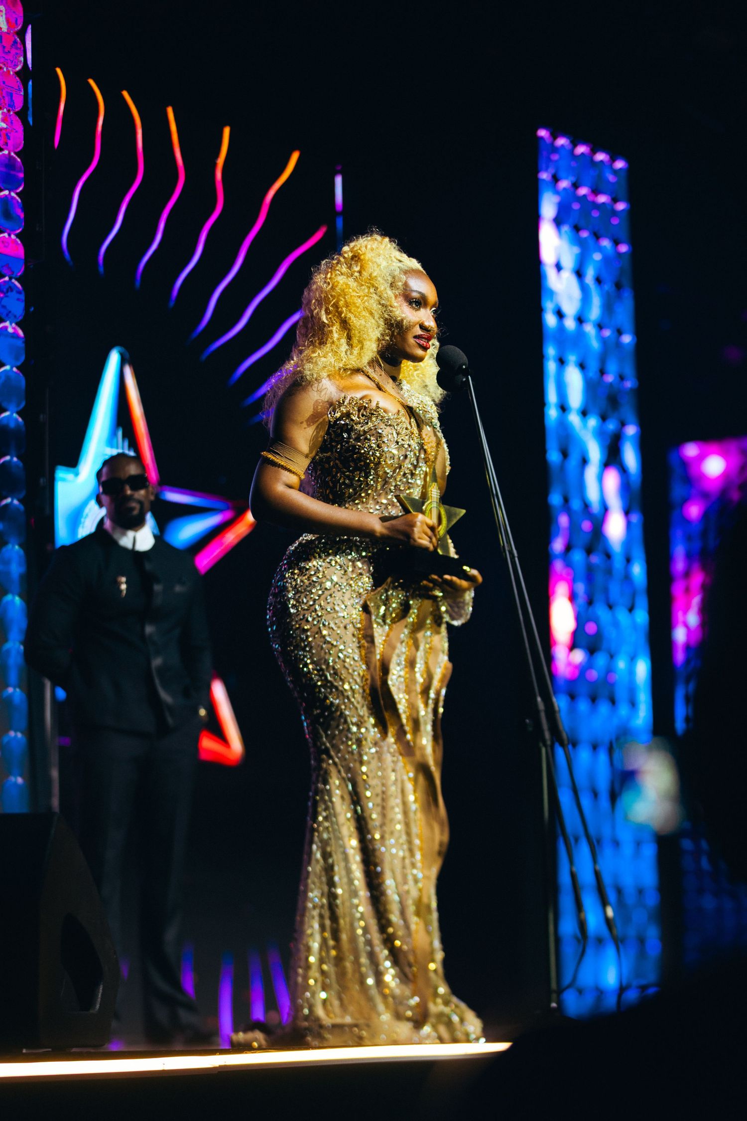 A woman in a stunning gold sequined gown accepts an award on stage at a glamorous ceremony.