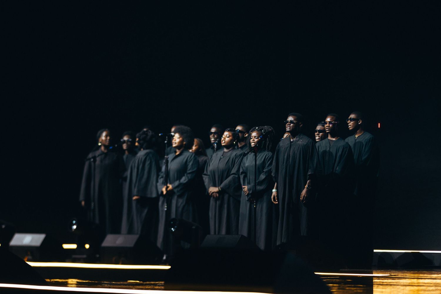 A gospel choir in dark robes performs on a dimly lit stage during a dramatic live concert.