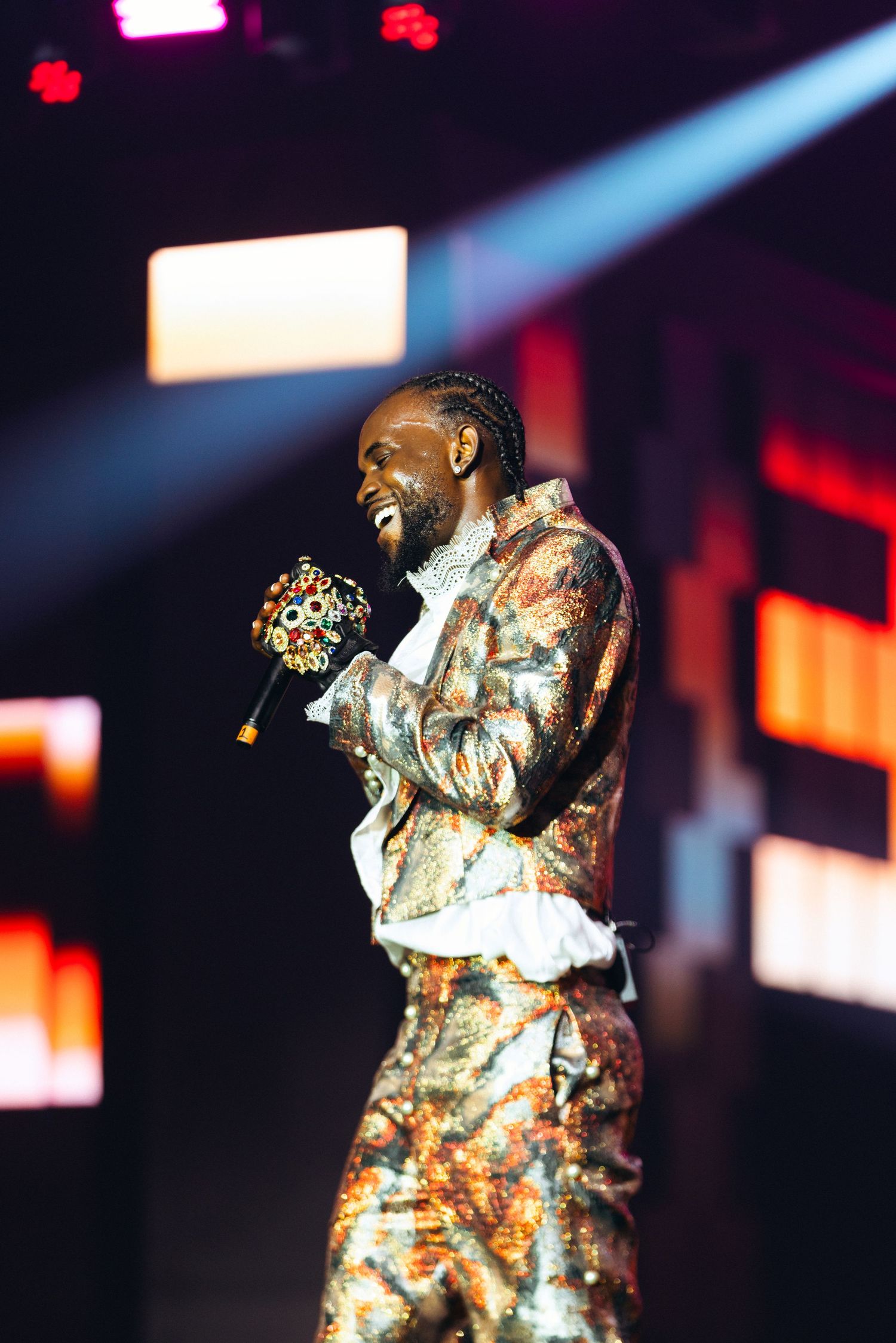 A performer in a gold patterned suit holds a decorative microphone on a colorful concert stage.