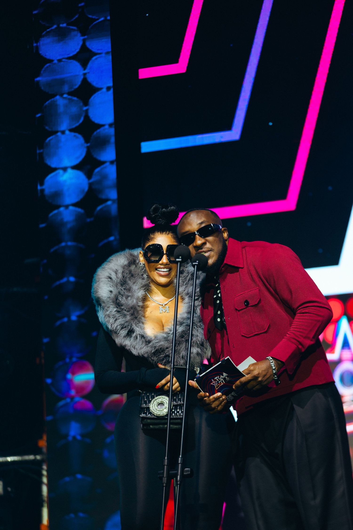 Two performers pose together on stage at an awards show, one wearing sunglasses and fur, the other in a red shirt.