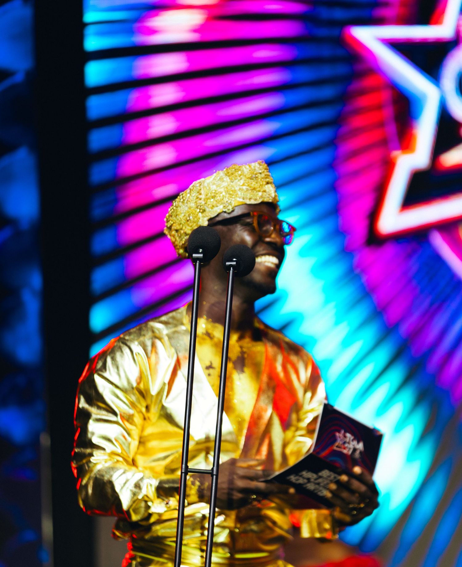 A man in a gold outfit and hat speaks at a microphone on a colorful neon-lit stage at an awards show.