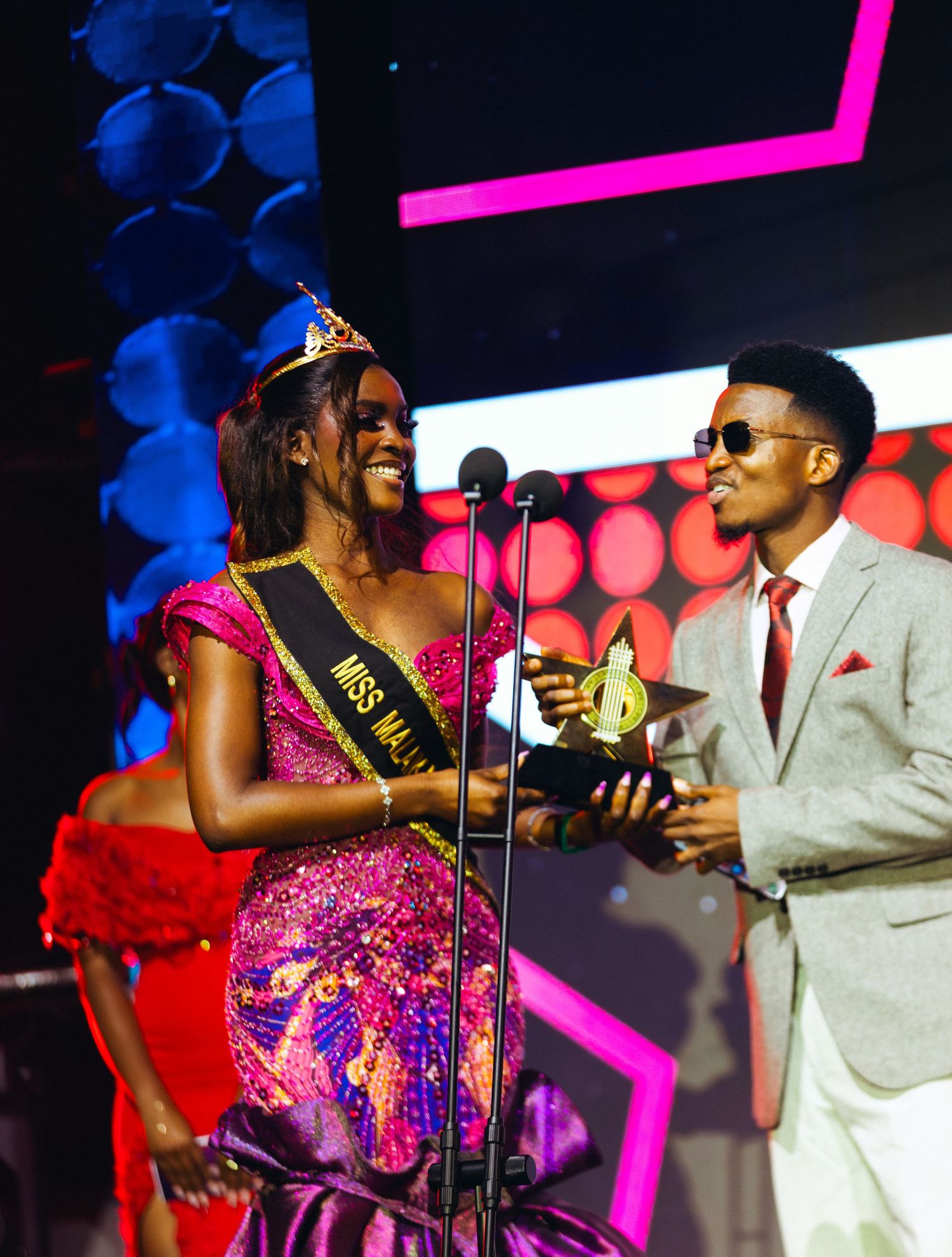 Miss Malawi beauty queen in pink sequin gown receives trophy on stage from man in grey suit.