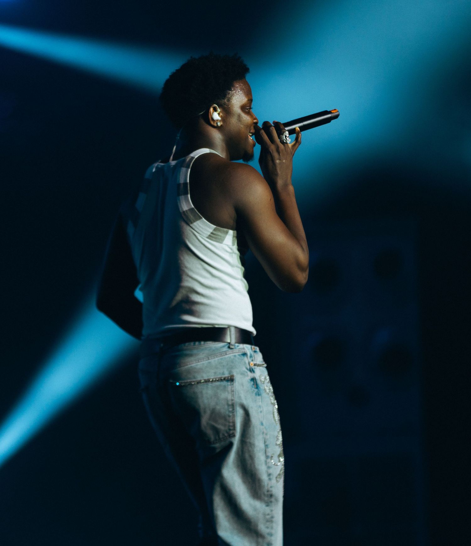 Young male performer singing into microphone on stage, wearing white tank top and jeans under blue concert lighting.