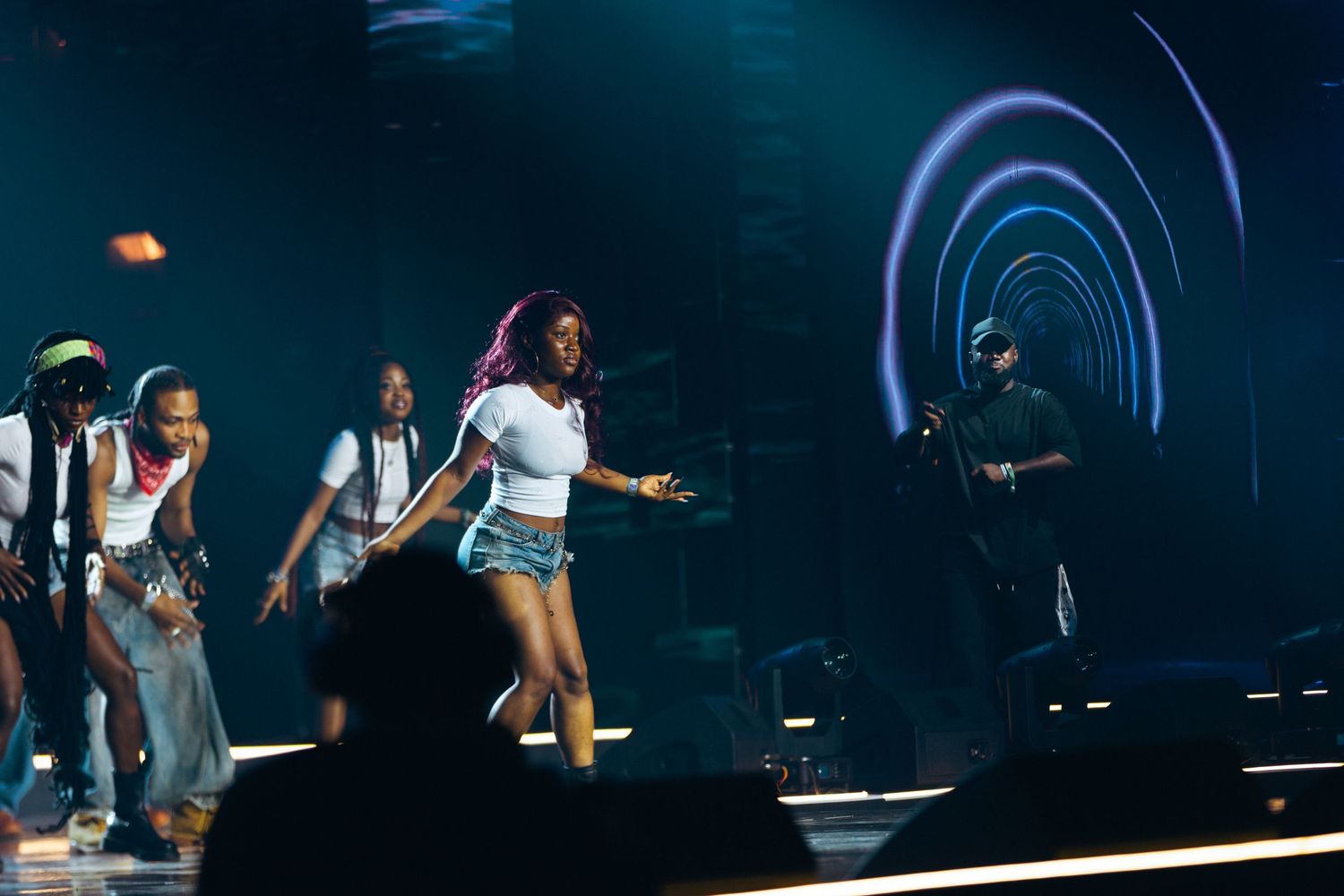 Female performer with pink hair in white top and denim shorts dancing on stage with backup dancers under blue lights.