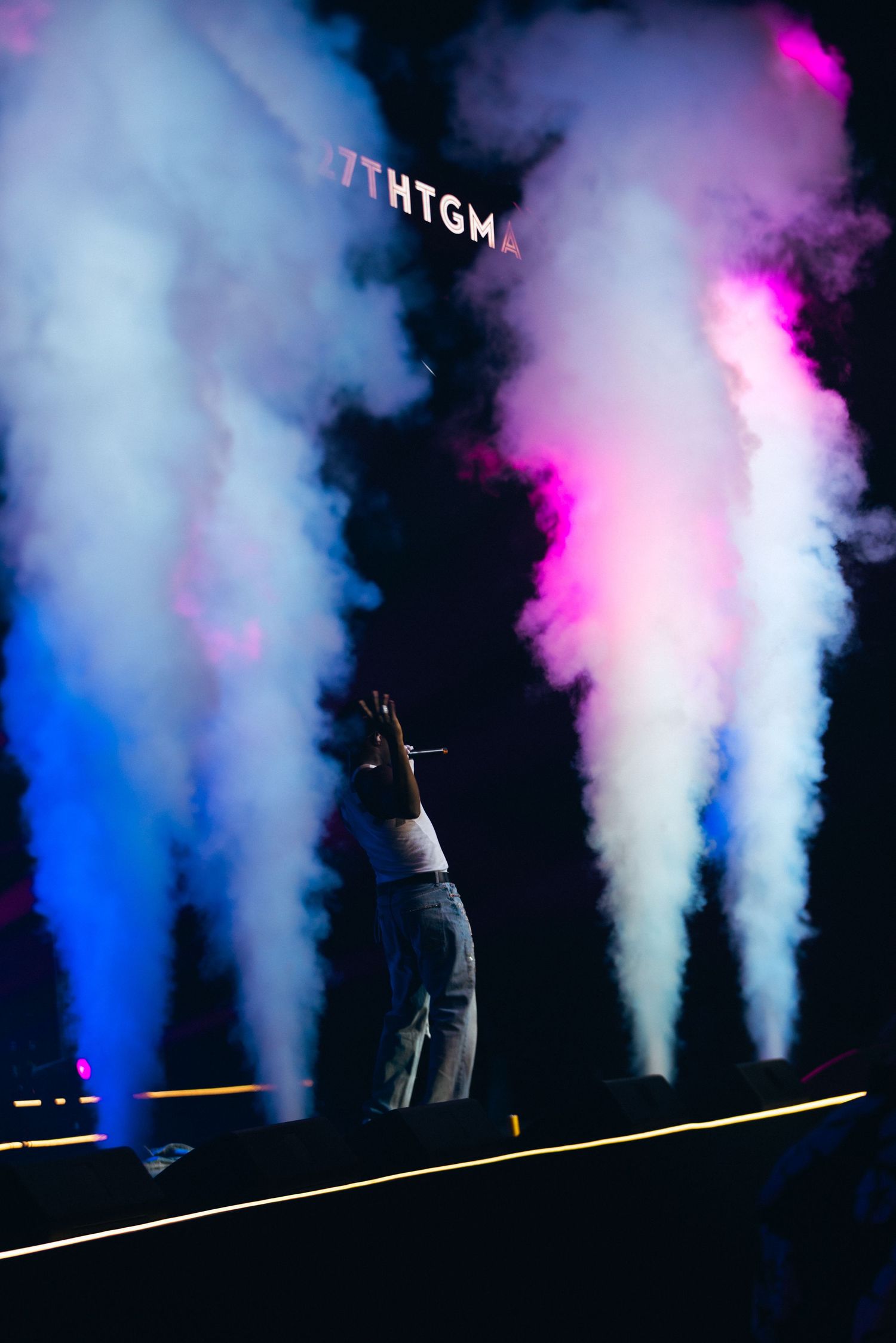 Performer on stage with colorful blue and pink smoke effects at a concert, raising hand with microphone.