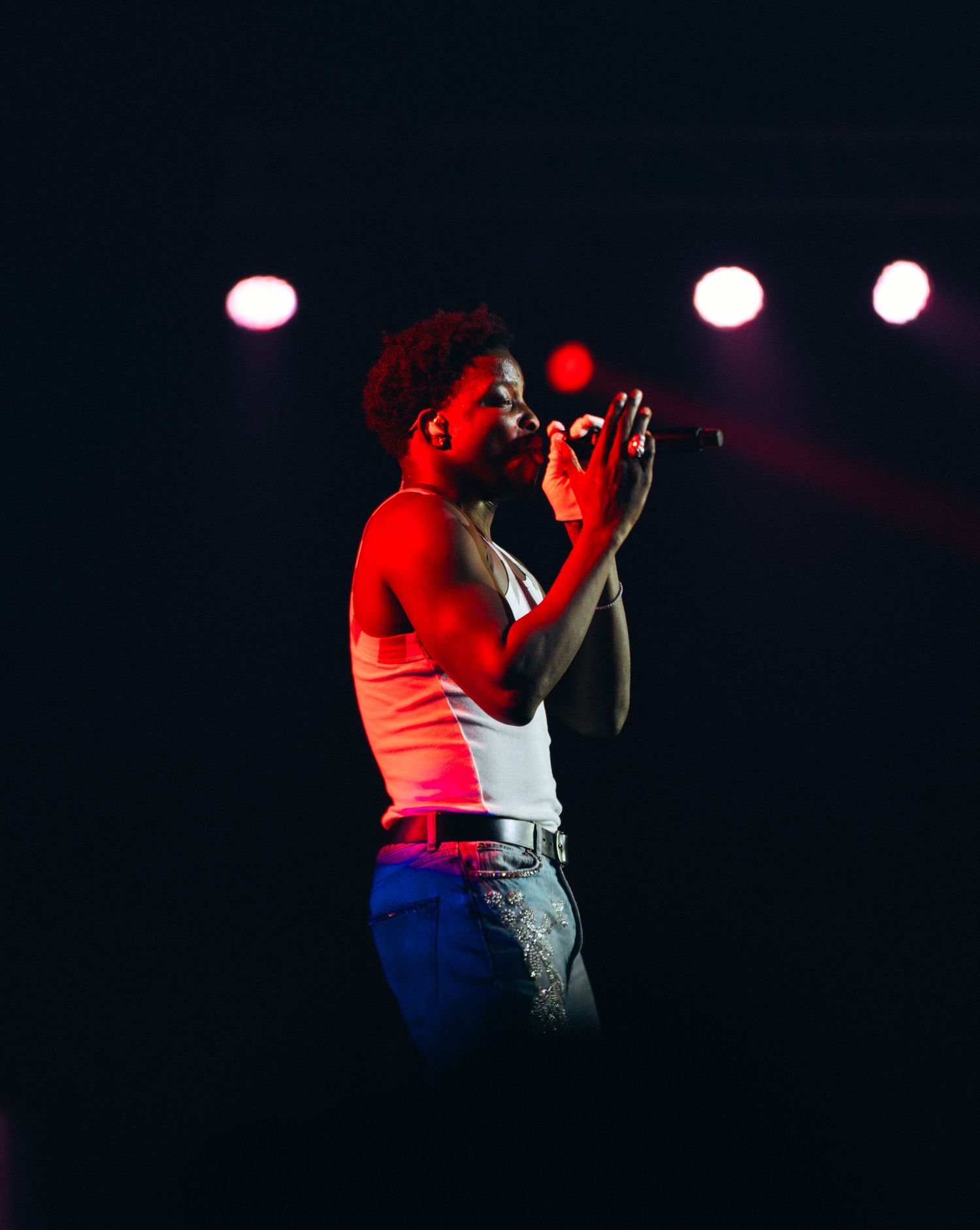 Male singer performing on stage under red stage lights, holding microphone while wearing white tank top and jeans.