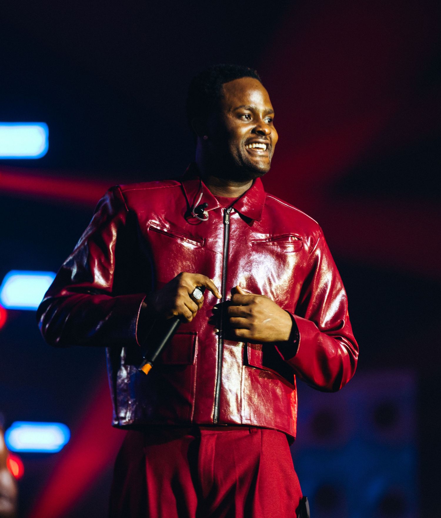 Man in red patent leather jacket smiling on stage holding microphone under colorful concert lights.