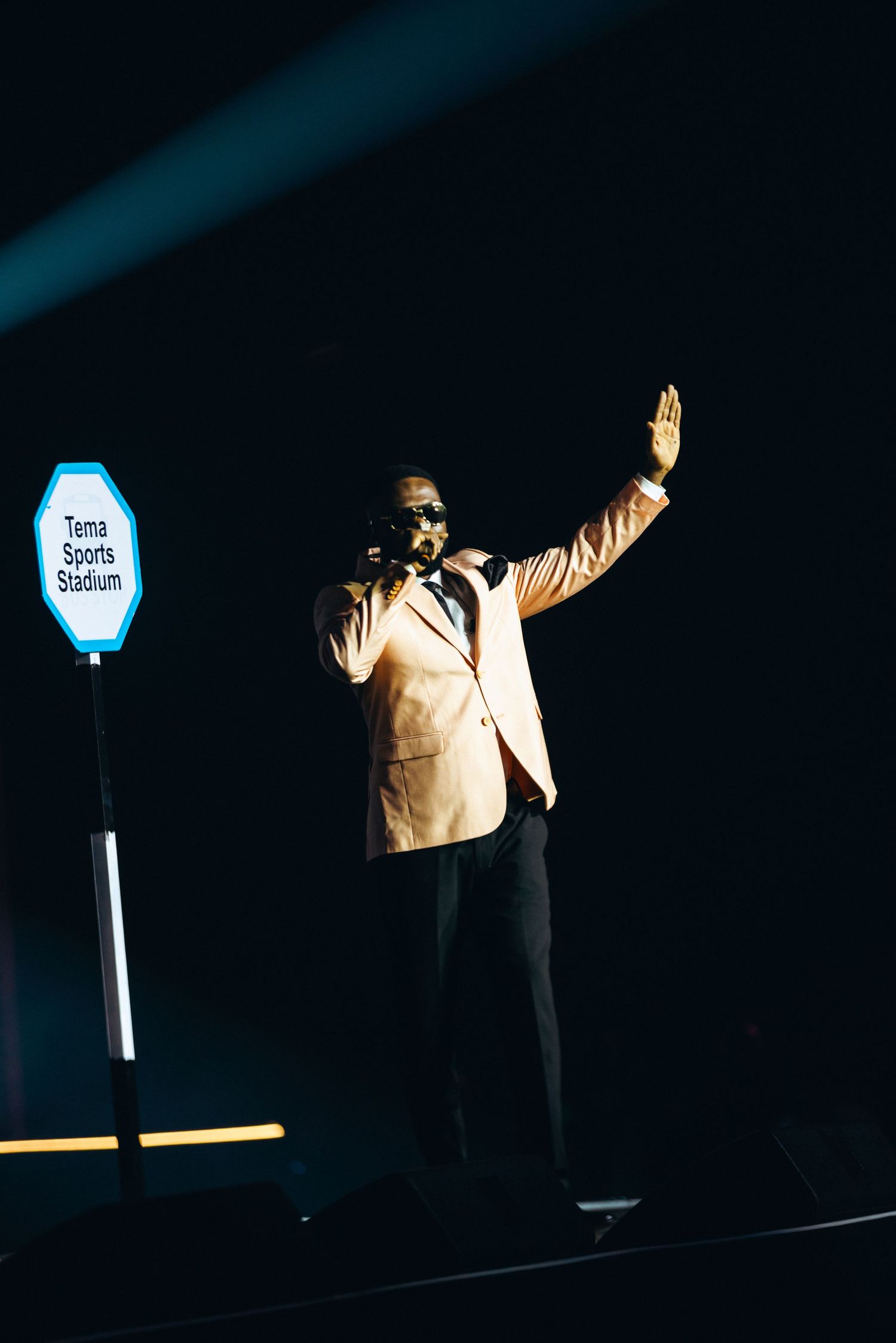 A male performer in a beige suit sings on a dark stage near a Tema Sports Stadium sign with hand raised.