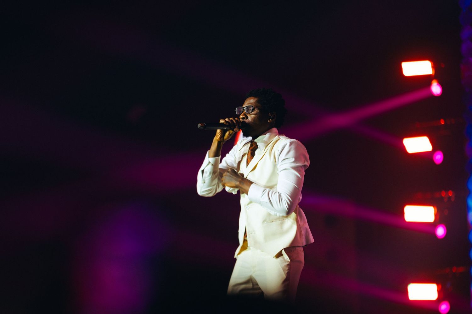 Male performer in white suit singing on stage with purple and pink stage lights behind him.